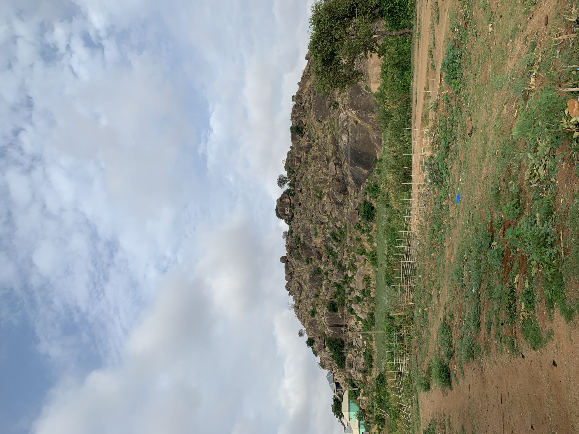 The image shows a rocky hill in Jos, Nigeria. The hillside is covered in sparse vegetation and dotted with large boulders. A rudimentary fence made of sticks runs along the base of the hill. The ground in front of the hill is a mix of bare earth and green foliage. In the lower left corner, there are some buildings partially visible. The sky is overcast with a mix of blue and white clouds.