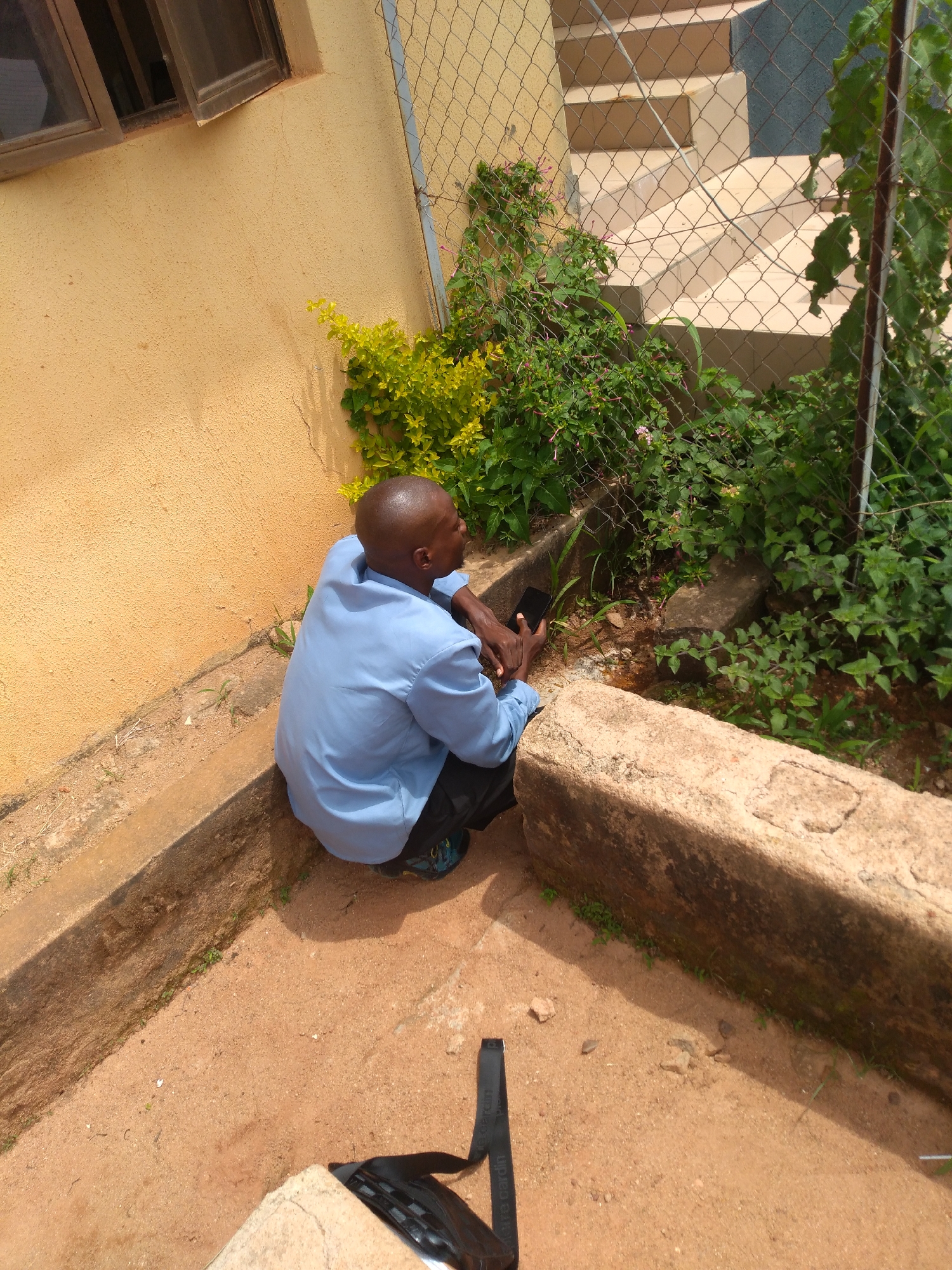 A man in a light blue collared shirt and dark pants is squatting on a concrete ledge next to a yellow building in an outdoor setting. He is holding a black smartphone in his right hand and appears to be looking at it. Behind him, a chain-link fence encloses a grassy area with several plants and a set of stairs. The ground is covered in reddish-brown dirt and small stones. The scene is located in Jos, Nigeria.