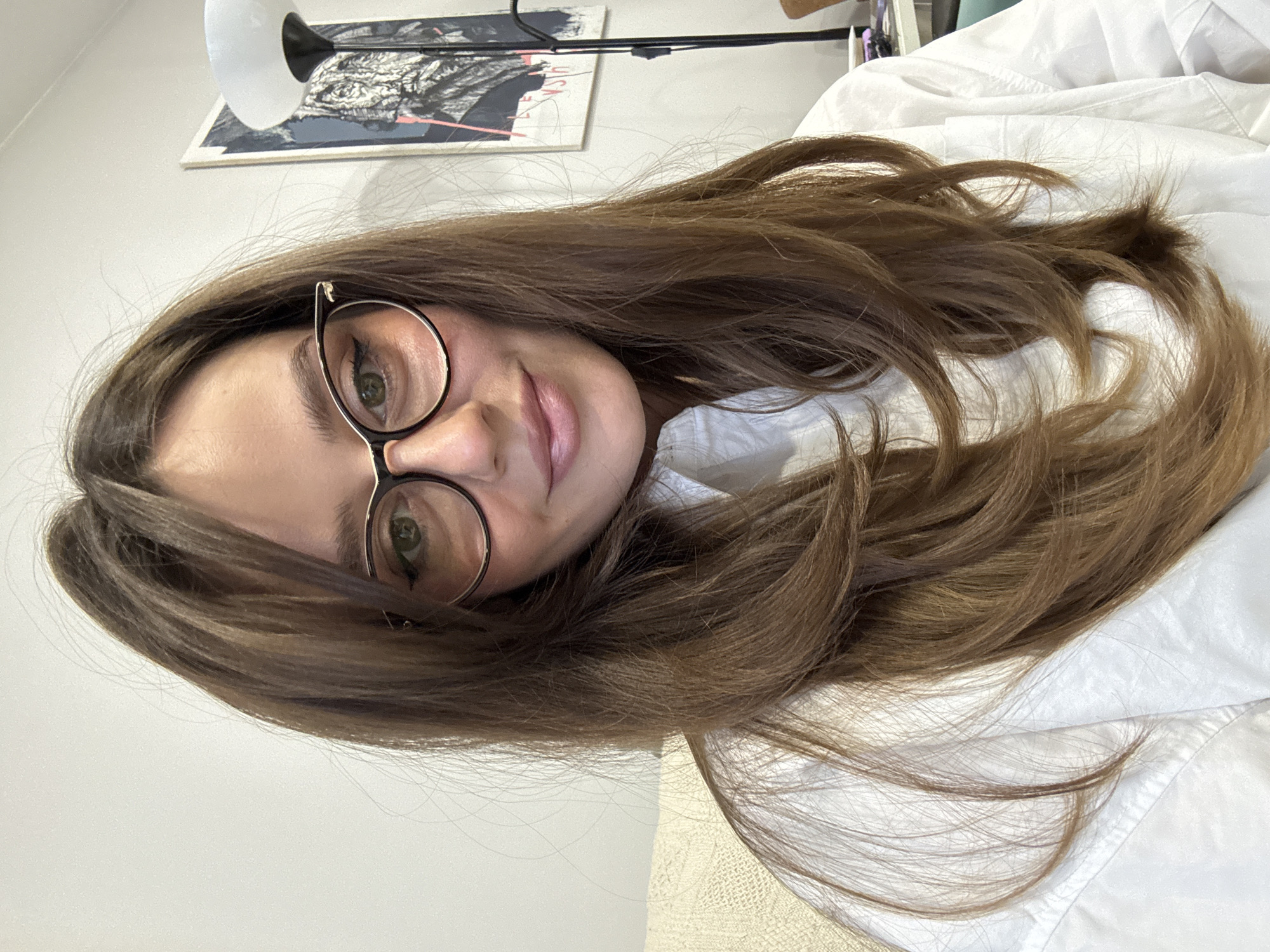 A woman with long, light brown hair and round glasses is lying down, facing the camera. She is wearing a white shirt. Behind her, a picture hangs on the wall and a desk lamp is visible. The location in Krasnoyarsk, Russia, is not discernible from the image.