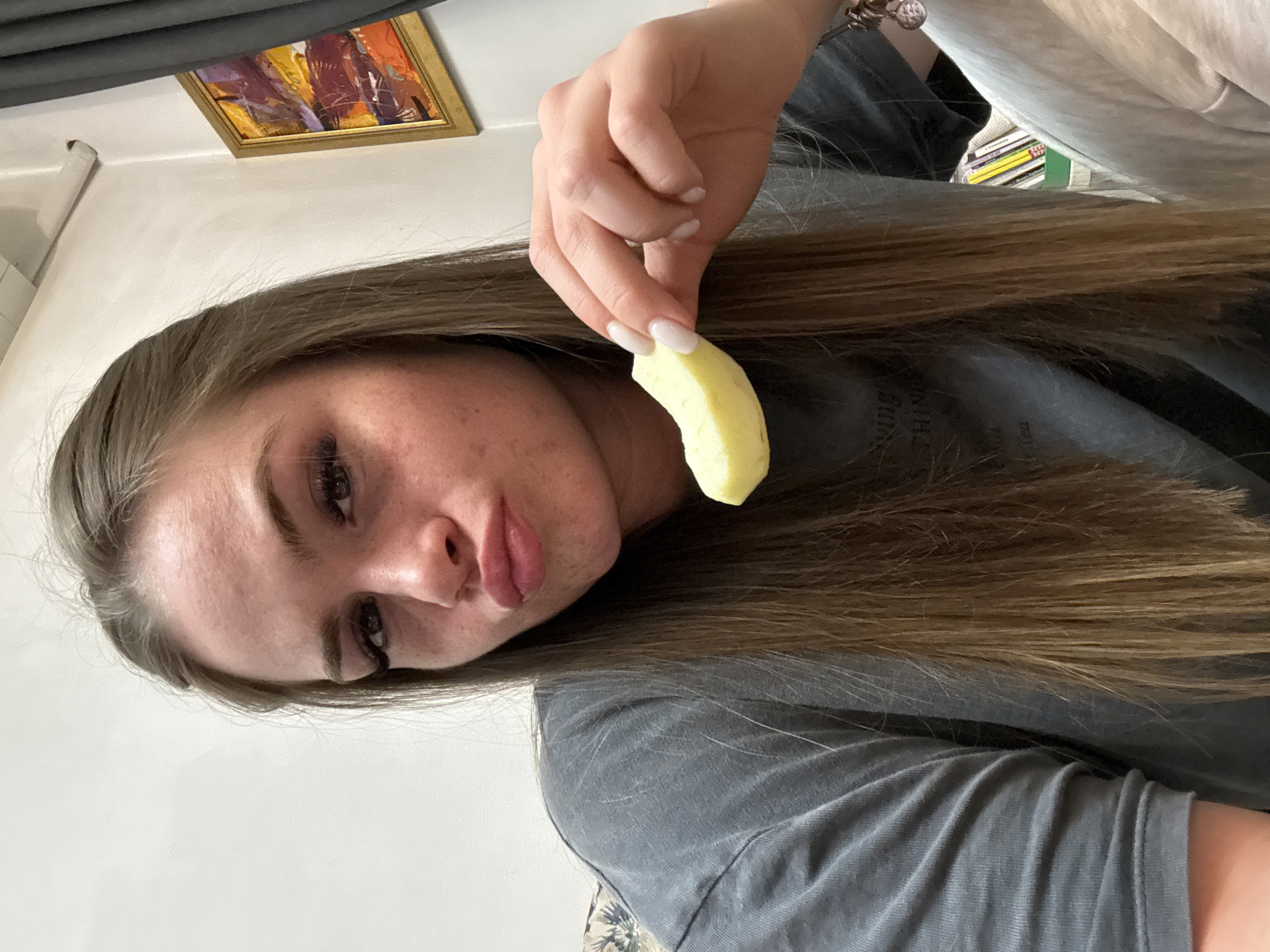 A woman with long brown hair is holding a slice of apple in her right hand. She is wearing a gray t-shirt. There is a framed painting on the wall in the background. The location is unknown and there is no information to indicate it is in Plovdiv, Bulgaria.