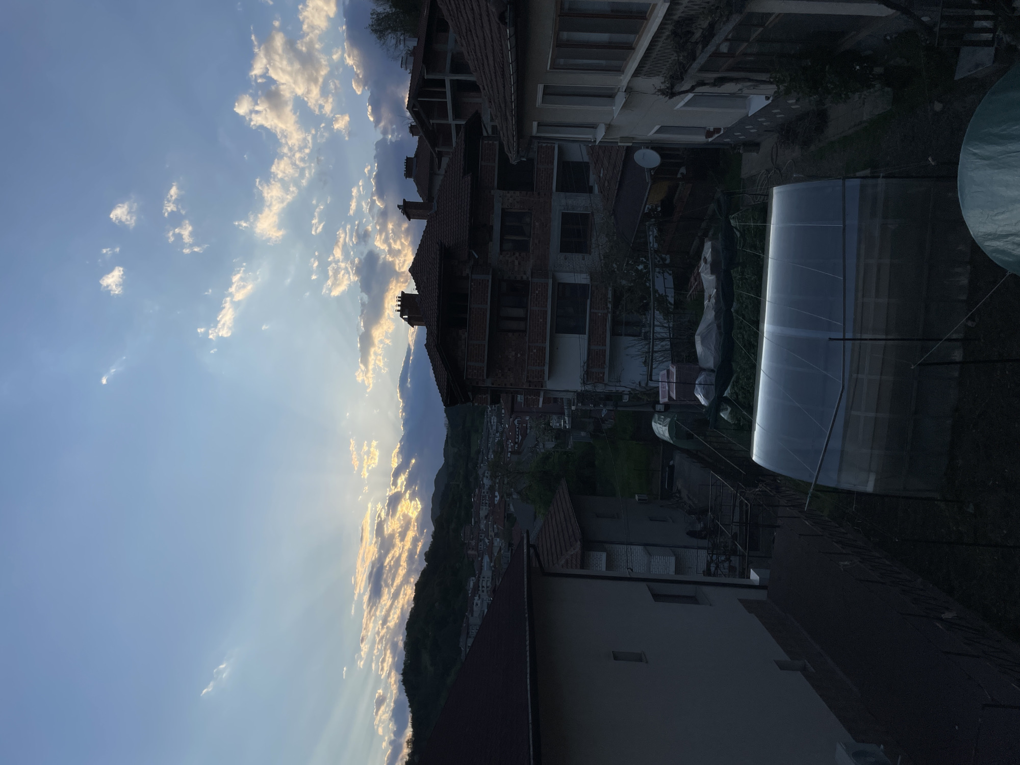 This image shows a view from a rooftop or elevated position in Zlatograd, Bulgaria, at dusk or dawn. The scene includes a neighborhood with houses, a forested hill in the background with rays of sunlight breaking through the clouds. There are buildings of varying heights and architectural styles, some with terracotta roofs, others with unfinished brickwork or siding. A plastic greenhouse is visible on the right side of the frame.