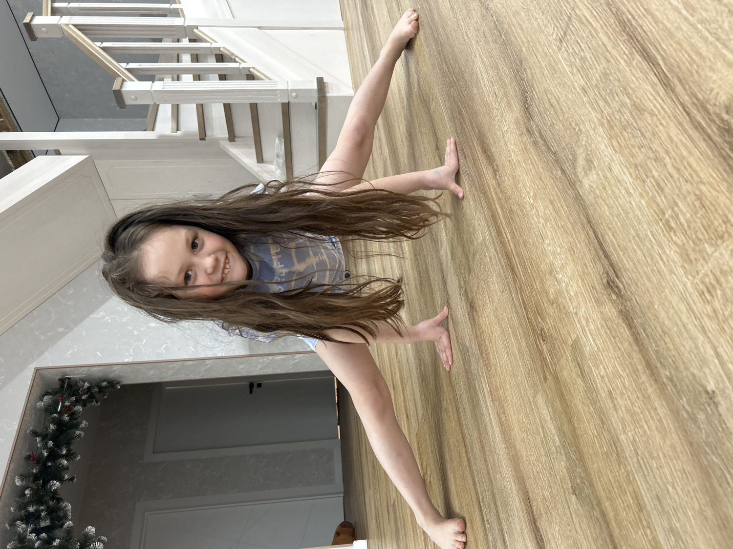 The image captures a young girl, approximately 6-10 years old, engaged in a playful pose indoors. She has long, dark brown hair that falls around her face and shoulders, and she is wearing a light-colored t-shirt with an indecipherable pattern or text on it. The girl is smiling broadly and looking directly at the camera, conveying a sense of happiness and cheerfulness.
She is positioned on a light brown, wood-patterned floor, performing a "crab walk" or bridge-like pose, with her hands and bare feet on the ground and her back arched. Her head is tilted, adding to the playful nature of the pose.
The setting appears to be a modern home in Chaikovsky, Russia. In the background to her left, a white staircase with decorative balusters and light wooden steps or handrails is visible, suggesting a multi-story residence. To her right and behind her head, white wall panels or cabinetry are present. Further to the far left, a festive Christmas garland with green foliage and hints of white lights can be seen, indicating the photo was likely taken during the winter holiday season. The interior is brightly lit, suggesting either ample natural light or effective artificial lighting, though the specific time of day is not discernible. The overall scene reflects a cheerful moment within a well-maintained home.