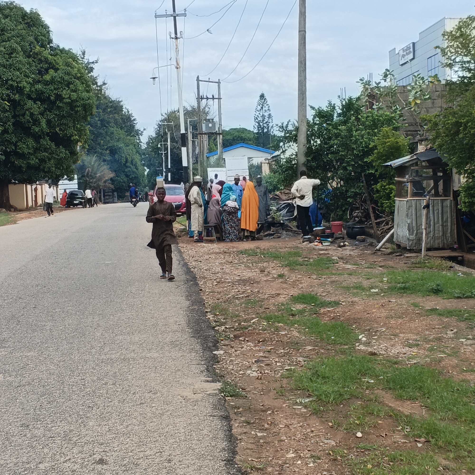 The image captures a street scene in Unguwar Rimi, Nigeria, with an overcast sky. A young boy in a dark traditional outfit is walking towards the camera on the right side of the asphalt road. In the distance, on the left side of the road, several people are gathered near parked vehicles and a small structure. Among them, a man in a white shirt and dark pants is standing, facing away from the camera, appearing to be involved in some activity. Further down the road, other individuals are visible, including a person on a motorcycle. The environment includes trees lining the left side of the road, and a patch of grass and dirt on the right, with a rudimentary outdoor shelter and cooking area. A modern building with signage is visible in the background on the right.