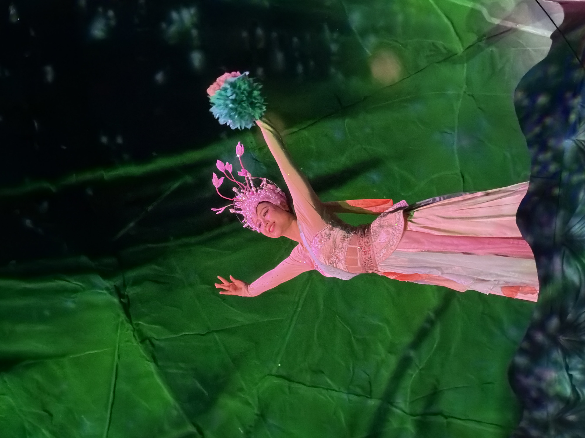 A female dancer performs on stage, dressed in an elaborate pink and white costume adorned with sequins and a jeweled headdress. She holds a fluffy green and pink pom-pom in her right hand and strikes a graceful pose with her arms extended. The backdrop is a large, textured green fabric, likely a theatrical drape, with subtle lighting effects creating a dappled pattern of light and shadow. The stage floor appears to be decorated with abstract, dark, and textured elements that blend into the shadows. The overall scene suggests a performance, possibly a cultural or theatrical production. The location is Unknown, Unknown.