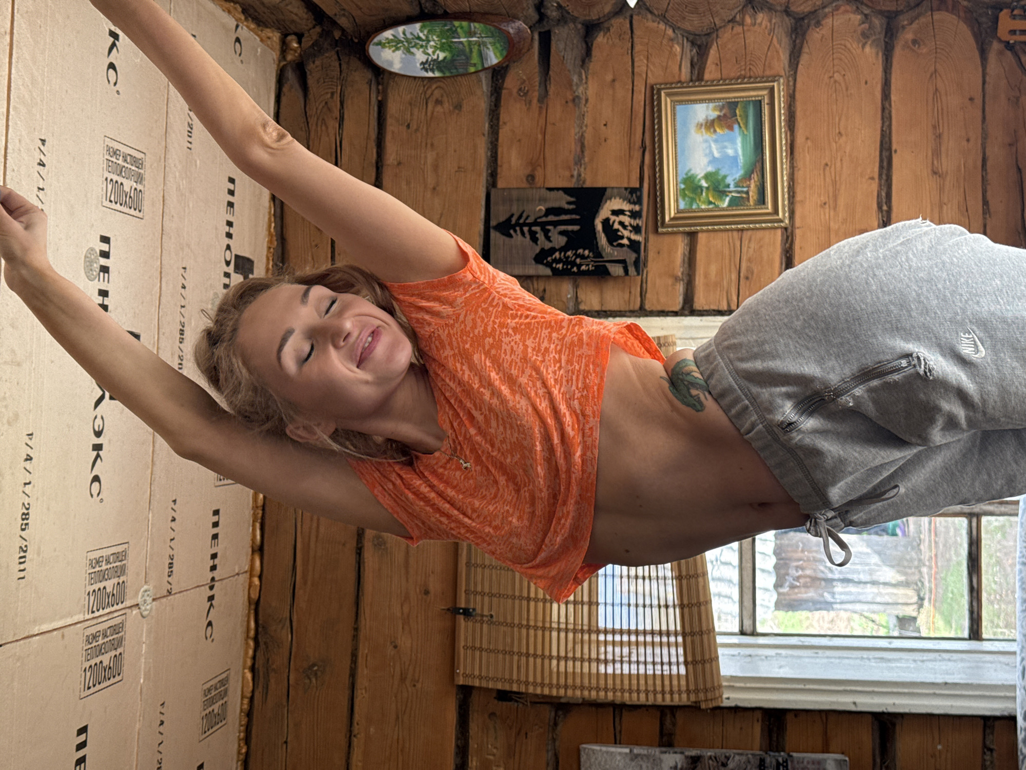 A woman is stretching indoors in a wood-paneled room. She is wearing a short orange shirt and grey sweatpants with a Nike logo. The walls of the room are lined with insulation panels. A window is visible behind her. There are several decorative items on the wall, including a small painting. The location is in Nizhnyaya Chermoda, Russia.