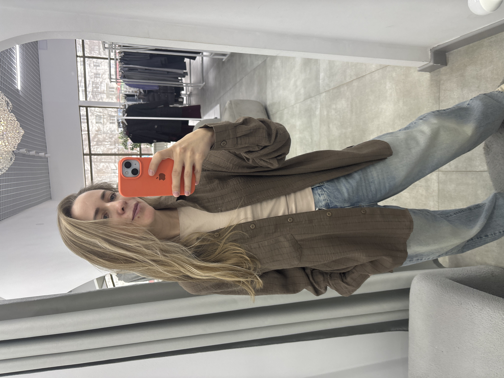 A woman is taking a selfie in a mirror. She has long, blonde hair and is wearing an open brown button-down shirt over a light-colored top and jeans. She is holding an orange iPhone in her right hand. The mirror is in what appears to be a clothing store, with racks of clothes visible in the background. The location is inferred to be in Chaikovsky, Russia, based on the context of the prompt. The environment is indoor, and the lighting appears to be artificial. There are no visible interactions or actions other than the woman taking the selfie.