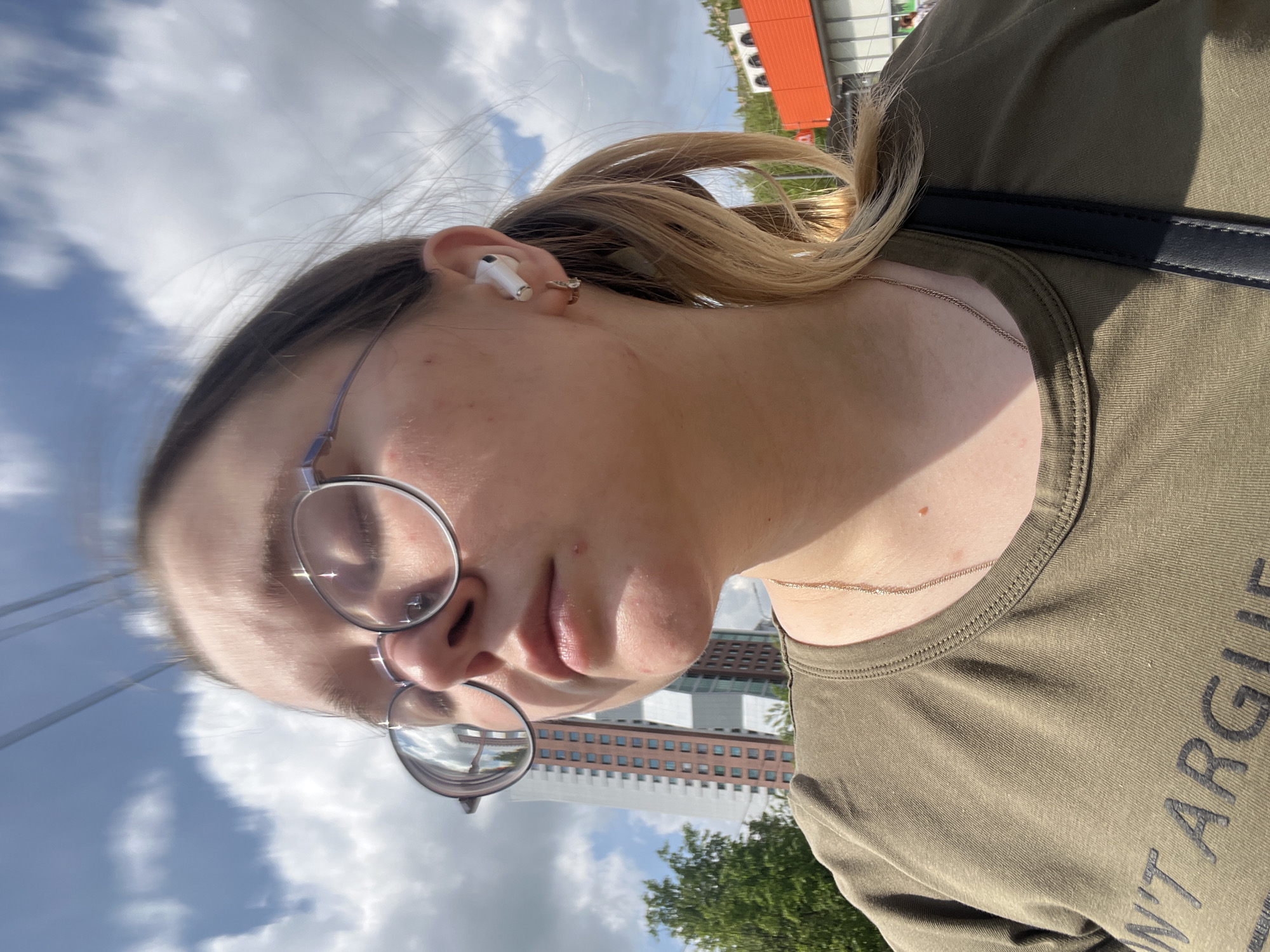 A close-up selfie of a woman wearing glasses, an AirPod in her ear, and an olive-green t-shirt with partial text reading "N'T ARGU". The background includes a cloudy sky, a building, and trees. The building with many windows is near Serpukhov, Russia.