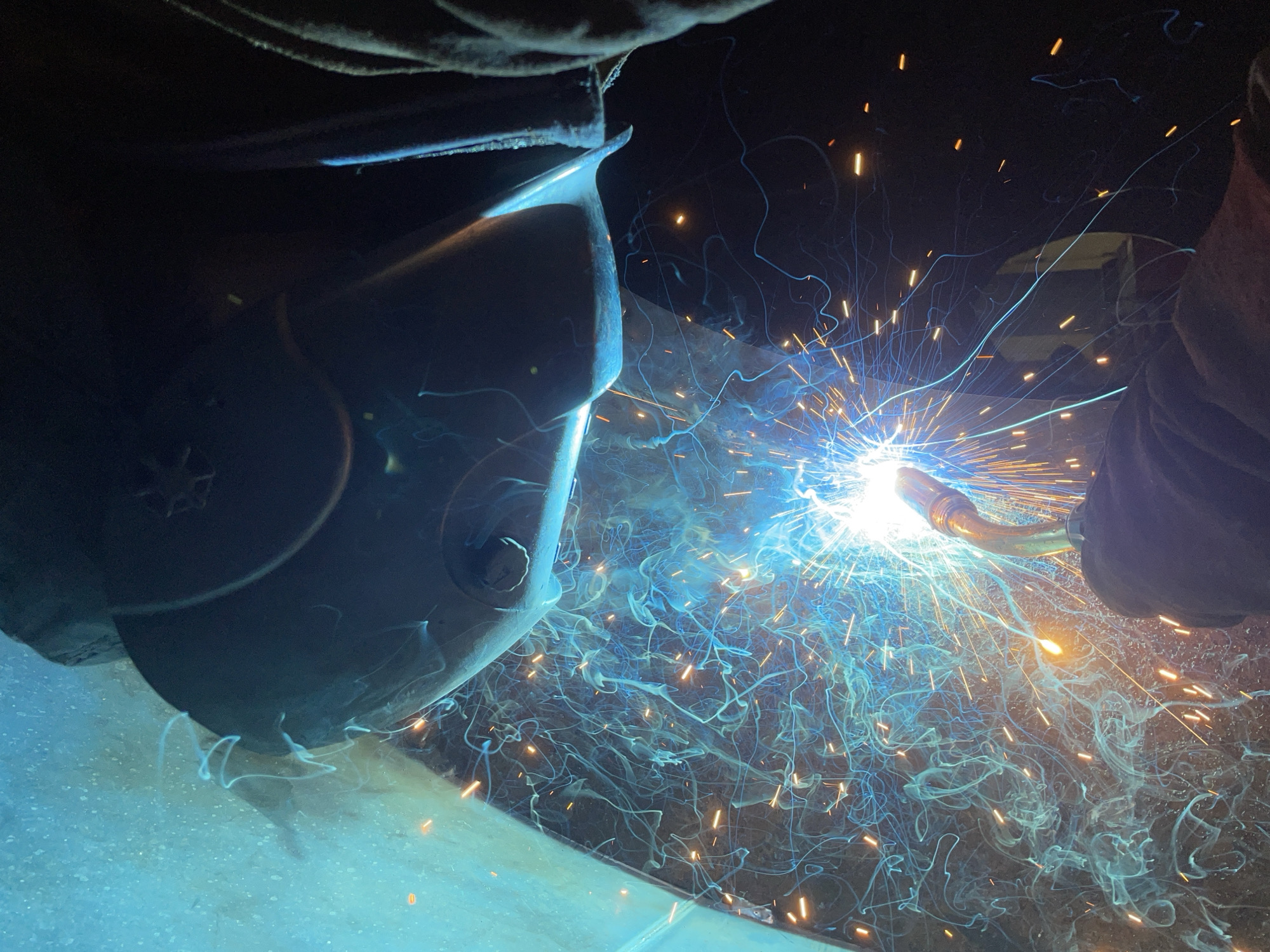 A person wearing a welding mask and gloves is welding metal. Sparks and fumes are emanating from the welding point. The location is unspecified and there is no indication that the scene is taking place in Čeluga, Montenegro.