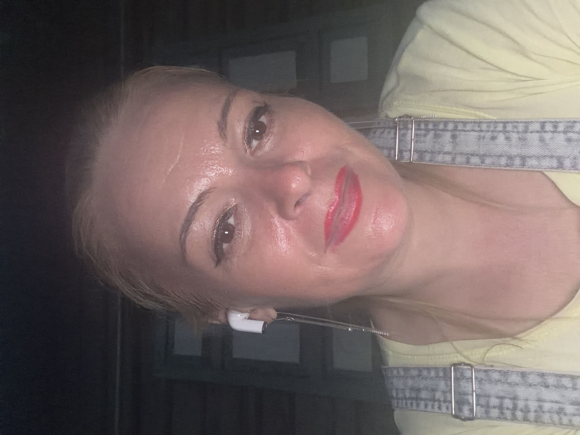 A woman is taking a selfie. She is wearing overalls over a yellow shirt. She has on red lipstick and what appears to be glitter eyeshadow. She is also wearing a white wireless earbud with a chain dangling from it. The background is dark and appears to be outdoors. The setting is unknown.