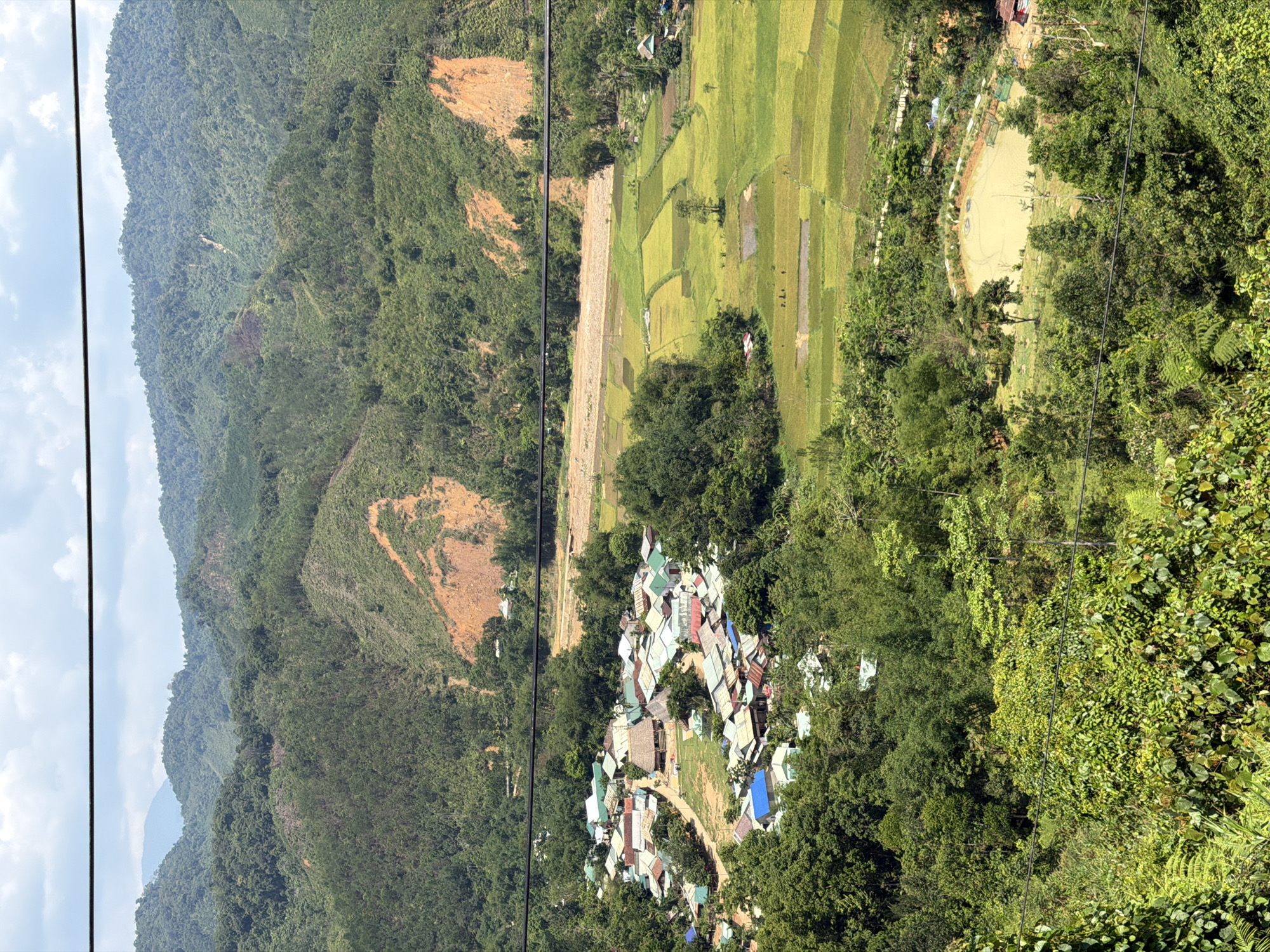 The image presents a high-angle view of a landscape, likely captured from a cable car or elevated position.
**Environment**: Dominated by lush, green mountainous terrain. A section of deforested land, exposing bare earth, is visible on the hillside. A patchwork of rice paddies stretches across a flatter area, displaying different stages of growth or harvest.
**Objects**: A cluster of buildings, possibly a small village or settlement, is nestled amidst the trees. These structures appear to have metal roofs, with variations in color. A circular body of water, potentially a pond or reservoir, is also visible. Cables from the cable car are seen in the top left part of the image.
**Location**: The location may be within the vicinity of Đà Nẵng, Vietnam, given the prevalence of rice paddies and mountainous terrain. However, more specific details would be required to verify.