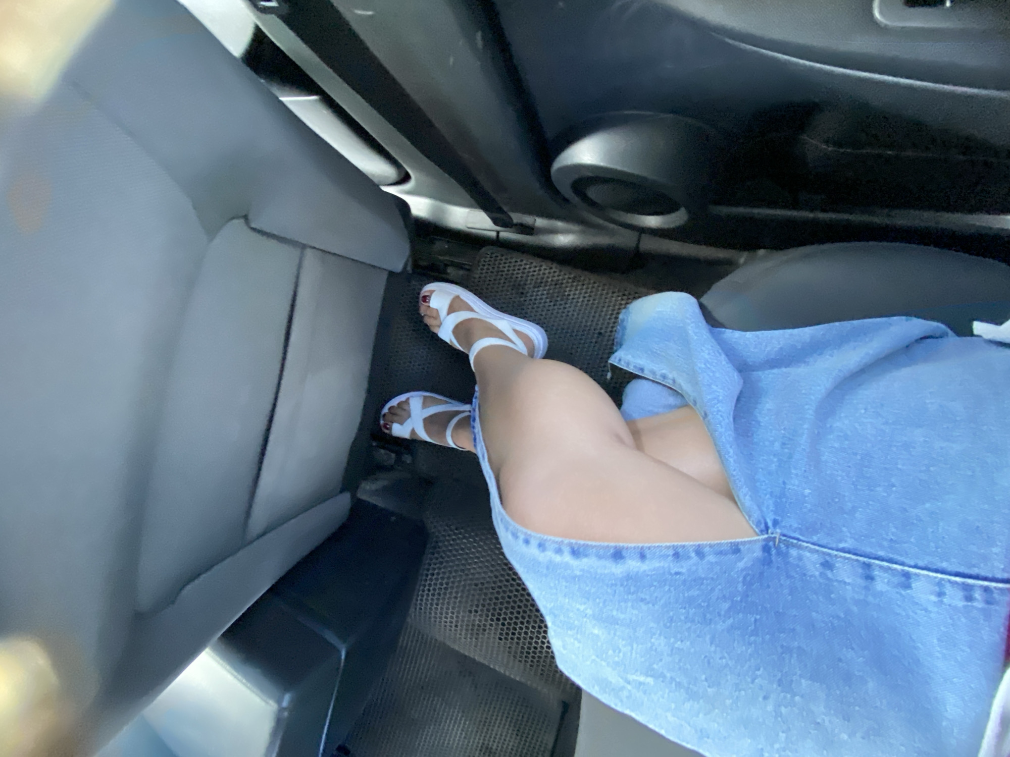 A person is sitting inside a vehicle. The person is wearing a denim skirt and white sandals. The interior of the vehicle is visible, including a seat and floor mat. The image appears to have been taken from a high angle, looking down at the person's legs and feet. The lighting suggests it is daytime. There is no information in the image to determine the location in the city of Zilair, Russia.