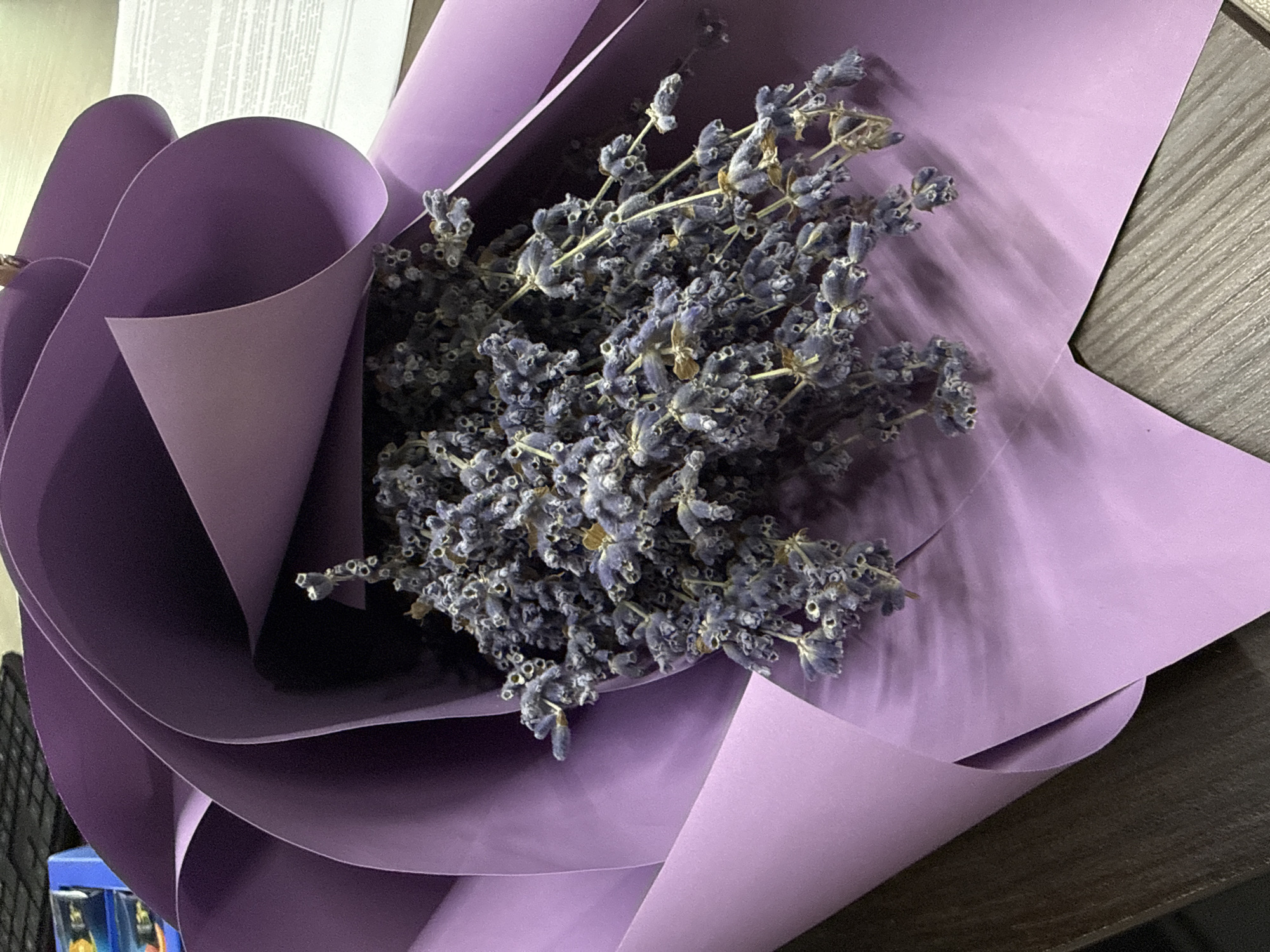 A close-up, overhead shot captures a bouquet of dried lavender flowers, predominantly in shades of purple and gray, nestled within a swirl of purple wrapping paper. The flowers are dry and delicate, with visible stems and individual florets. The wrapping paper is a muted, light purple, creating a soft backdrop and framing the lavender. Shadows are cast across the paper, suggesting a light source from the upper left. The bouquet rests on a dark, textured surface, possibly a wooden table. In the background, a portion of a keyboard and a blue object are visible. The scene is devoid of people or any discernible activity. No information regarding a specific location in Nizhny Novgorod, Russia, is evident from the image itself.