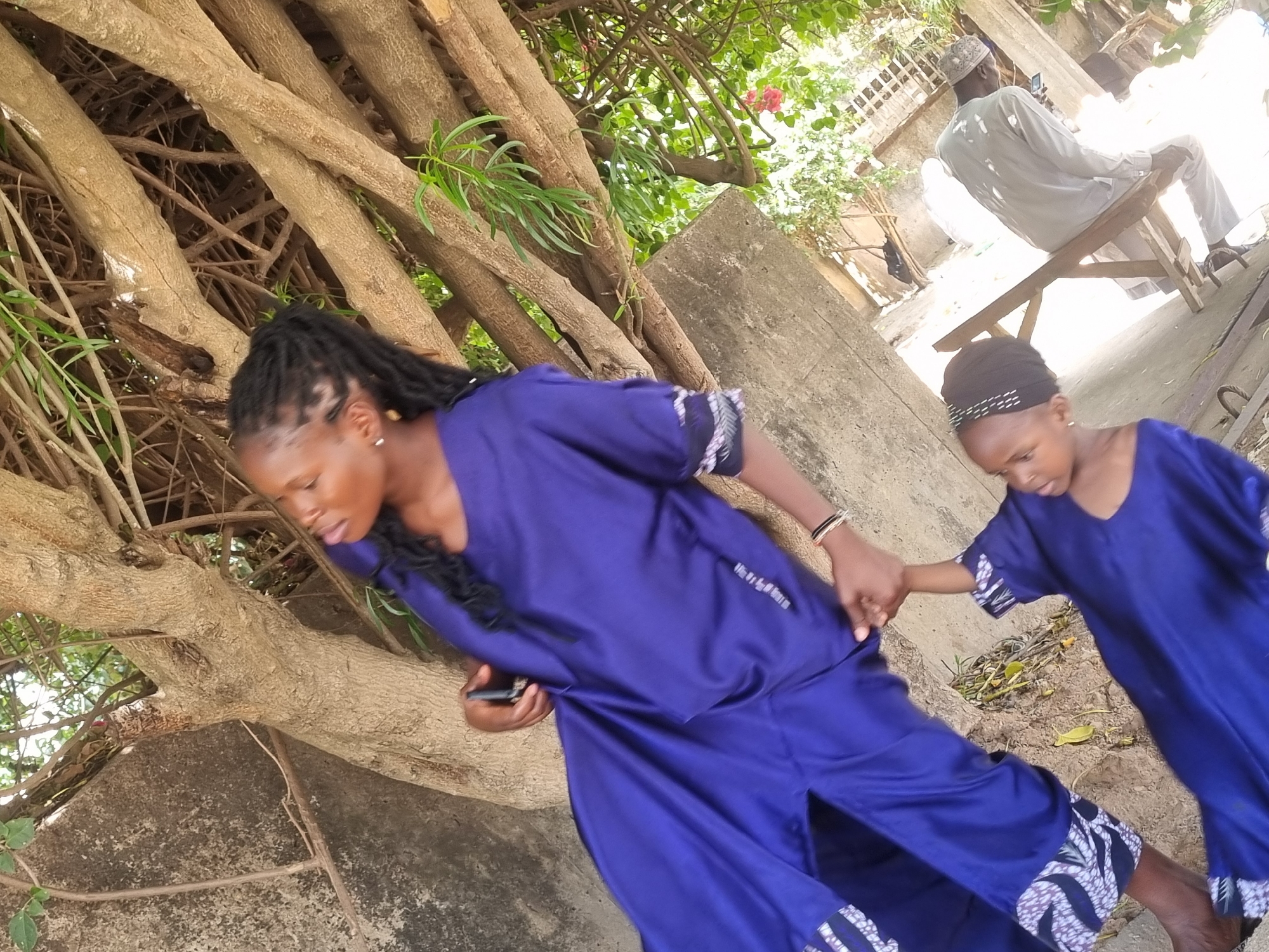 The image depicts a woman and a child, likely female, both wearing matching blue dresses with patterned borders. The woman has dreadlocks and is holding a mobile phone. She is holding the child's hand. They are walking through a park-like area in Jos, Nigeria, with tree branches overhead and a concrete wall or structure to their left. In the background, a man is sitting on a wooden bench, also appearing to be using a mobile phone. The environment suggests a moderately sunny day.