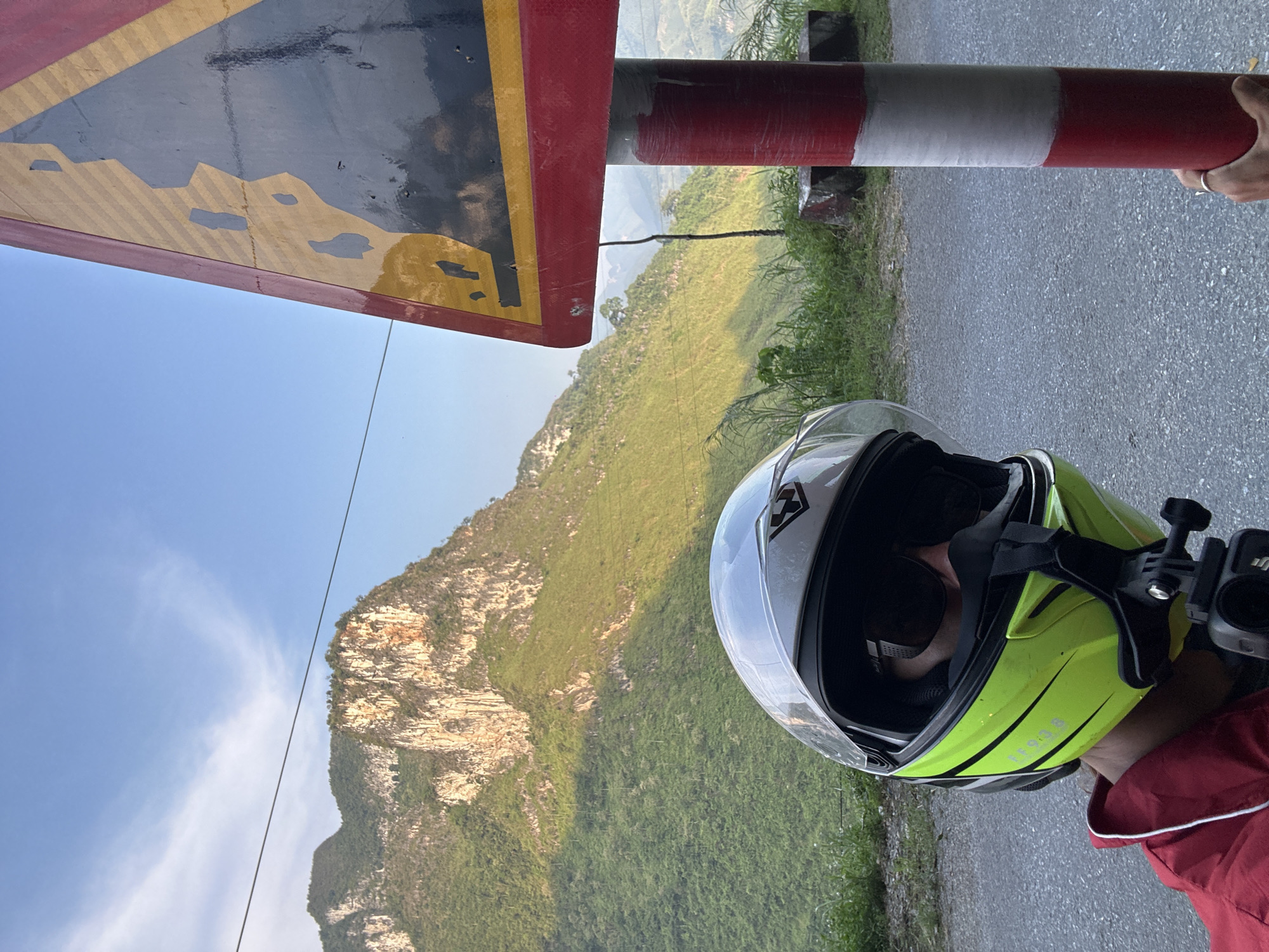 A person wearing a yellow motorcycle helmet with a clear visor stands on a paved road in Đường Thượng Commune, Vietnam. The road is adjacent to a lush, green mountain terrain. A red-and-white striped cylindrical barrier is on the right, near the person. A triangular sign with a black-and-yellow hazard warning is partially visible at the top left. The sky is clear blue.