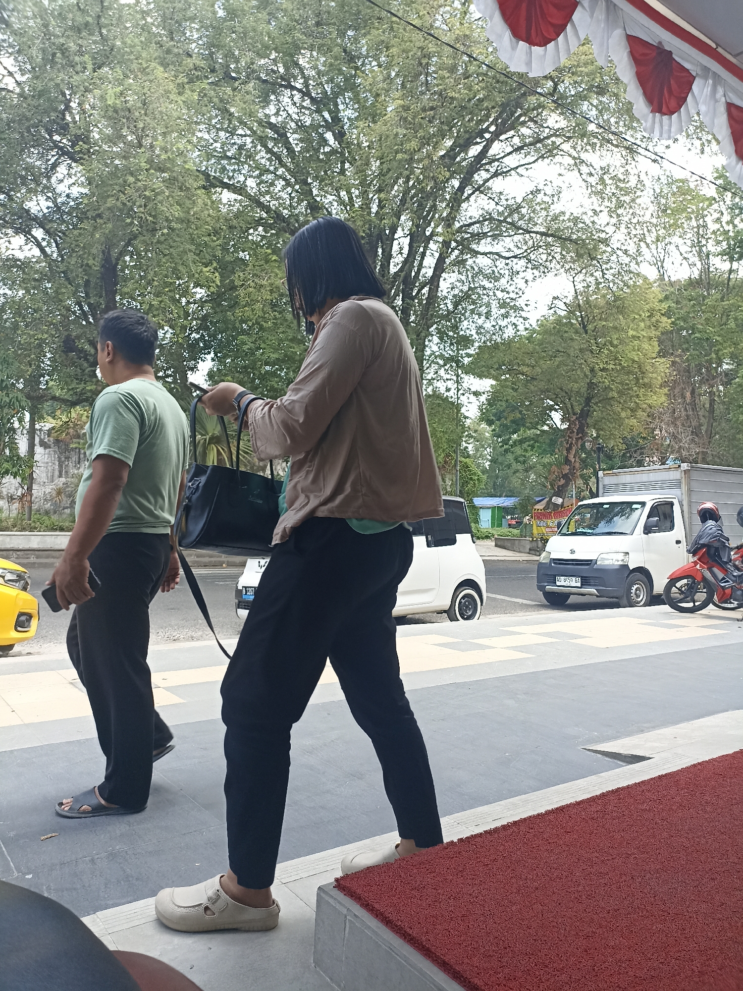 In Surakarta, Indonesia, two individuals are walking outdoors near a street. The person on the left, wearing a green t-shirt and black pants, carries a mobile phone in their right hand. The person on the right, with long dark hair, wears a light brown long-sleeved shirt and black pants, and carries a dark handbag with their left hand while looking at their phone. A white car and a white truck are visible on the street, along with a red motorcycle. The environment includes trees and decorative red and white banners overhead. The ground is paved with tiles and a red mat is visible.