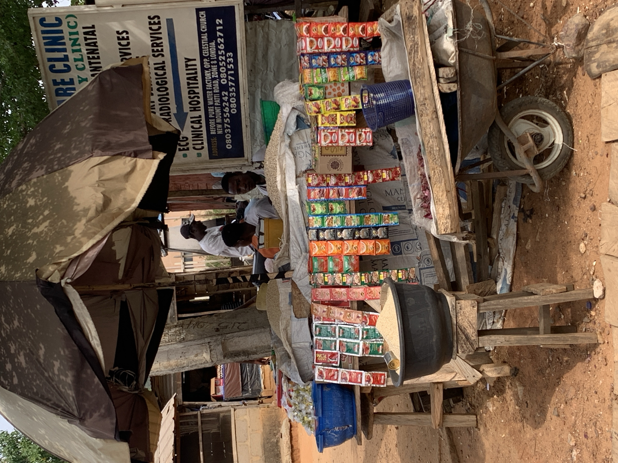 The image depicts a street scene in Lokoja, Nigeria. There's an outdoor stall set up under an umbrella and a sign for "RE Clinic (Y Clinic)" and "RADIOLOGICAL SERVICES". The stall is filled with packaged snacks. Behind the stall, two people are visible, both wearing white shirts. Next to the stall is a wheelbarrow. The address on the sign indicates the location is "BEFORE PURE WATER FACTORY, OPP. CELESTIAL CHURCH, NEW MOUNT PATTI ROAD, ZONE 8 LOKOJA". A black container of some sort (possibly containing rice or similar) is also visible. The setting appears to be a somewhat informal and possibly improvised business setup.