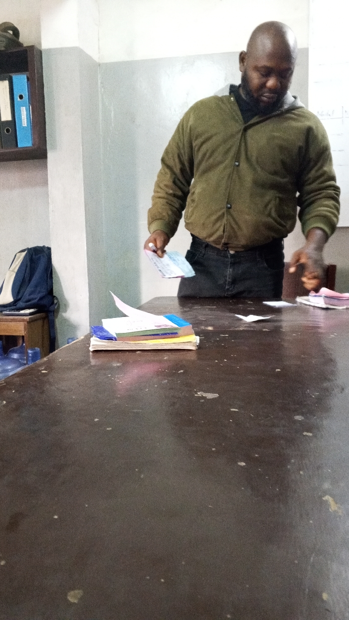An indoor scene in Dakata, Nigeria, depicts a dark-skinned male subject, bald with a short beard, standing behind a dark, worn wooden table. He wears an olive green hooded jacket over a black shirt and dark jeans. His gaze is directed downwards towards the table surface. In his left hand, he holds a light-colored document, while his right hand is extended over the table towards a small stack of pink-bound items. A prominent stack of multi-colored notebooks or papers with some bent pages rests on the table's left side. The table surface exhibits marks and wear. In the background to the left, a wooden shelf holds black, white, and blue binders. Below the shelf, a dark blue and light blue backpack sits on a small wooden stool, with a blue plastic container visible on the floor. The walls are painted white and light gray, showing signs of wear. A faint whiteboard with illegible writing is partially visible in the upper right background.