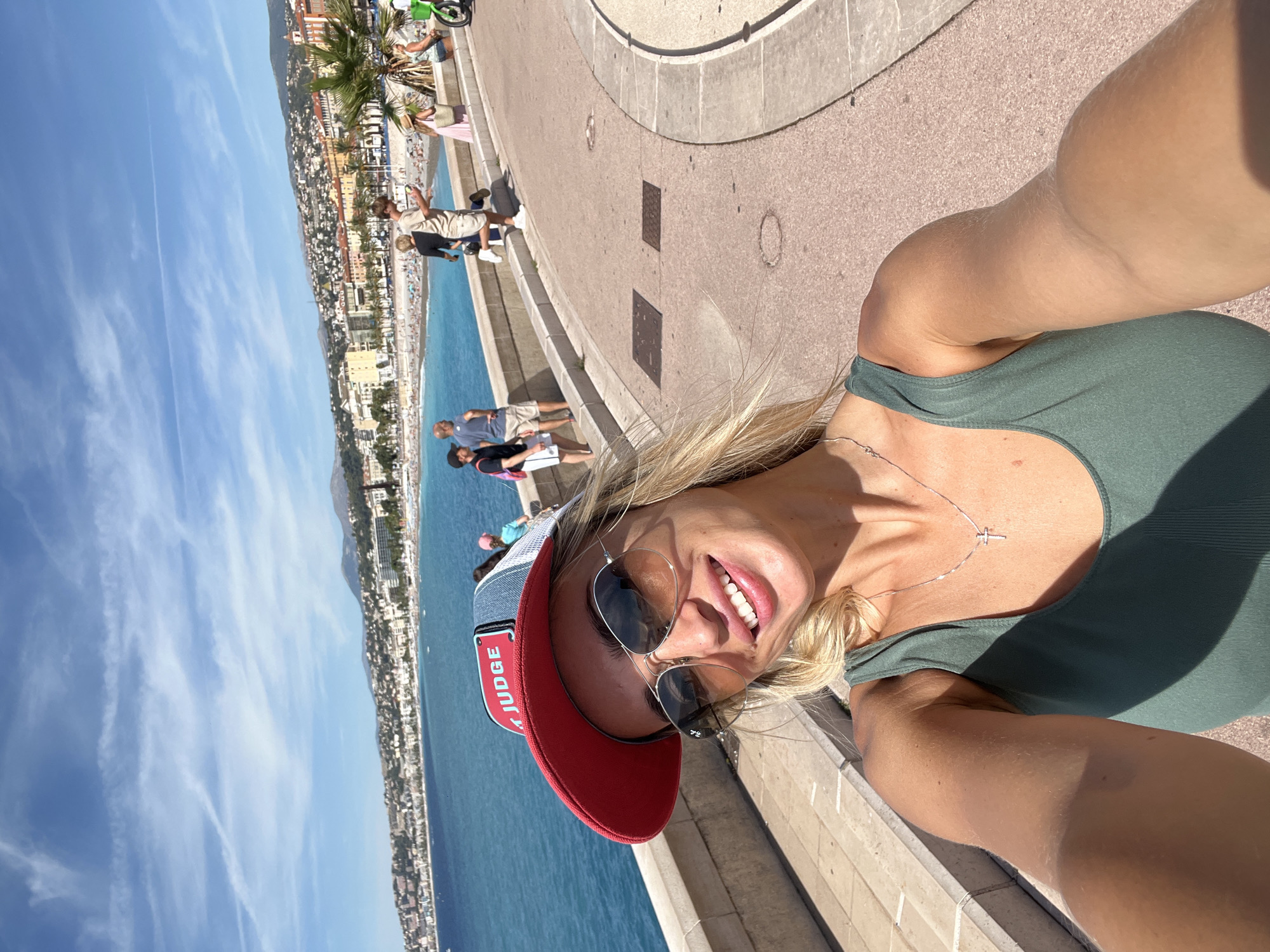 A selfie is taken by a blonde woman with sunglasses and a red and blue baseball cap, showing her smiling at the camera. She is wearing a green tank top and a silver cross necklace. In the background, the coastline of Nice, France, is visible, with a blue sea, a beach dotted with people, and a city with buildings stretching along the shore. A promenade with a paved walkway and a low stone wall separates the foreground from the beach. Several people are walking on the promenade, and a green electric scooter is parked to the right. The sky is blue with some wispy clouds.