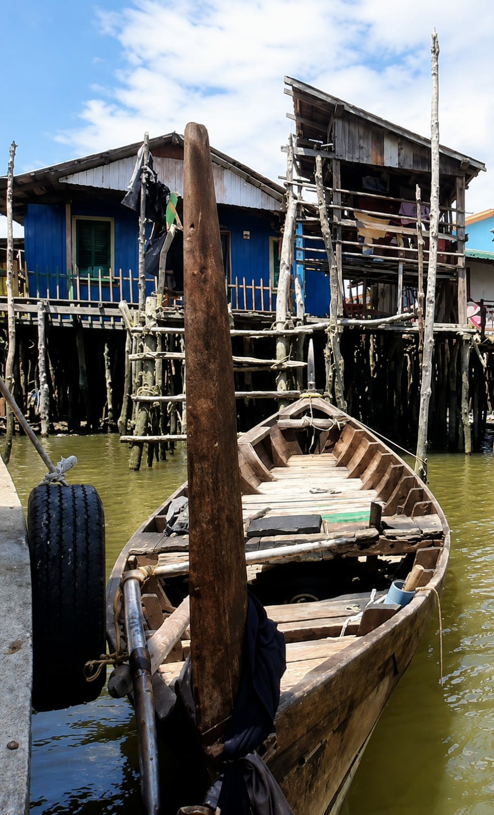 A weathered wooden boat is docked next to a concrete pier in front of stilted, weathered wooden structures. The boat is empty and appears to be in a state of disrepair, with visible cracks and missing planks. A large tire is attached to the pier, acting as a fender. The background features two stilted buildings, one painted bright blue and the other a natural wood color. Both structures exhibit signs of age and wear. The water is murky green, and the sky is partially cloudy. The scene appears to be a waterfront area, possibly a fishing village or a residential area built on stilts over water, in the city of Medan, Indonesia.