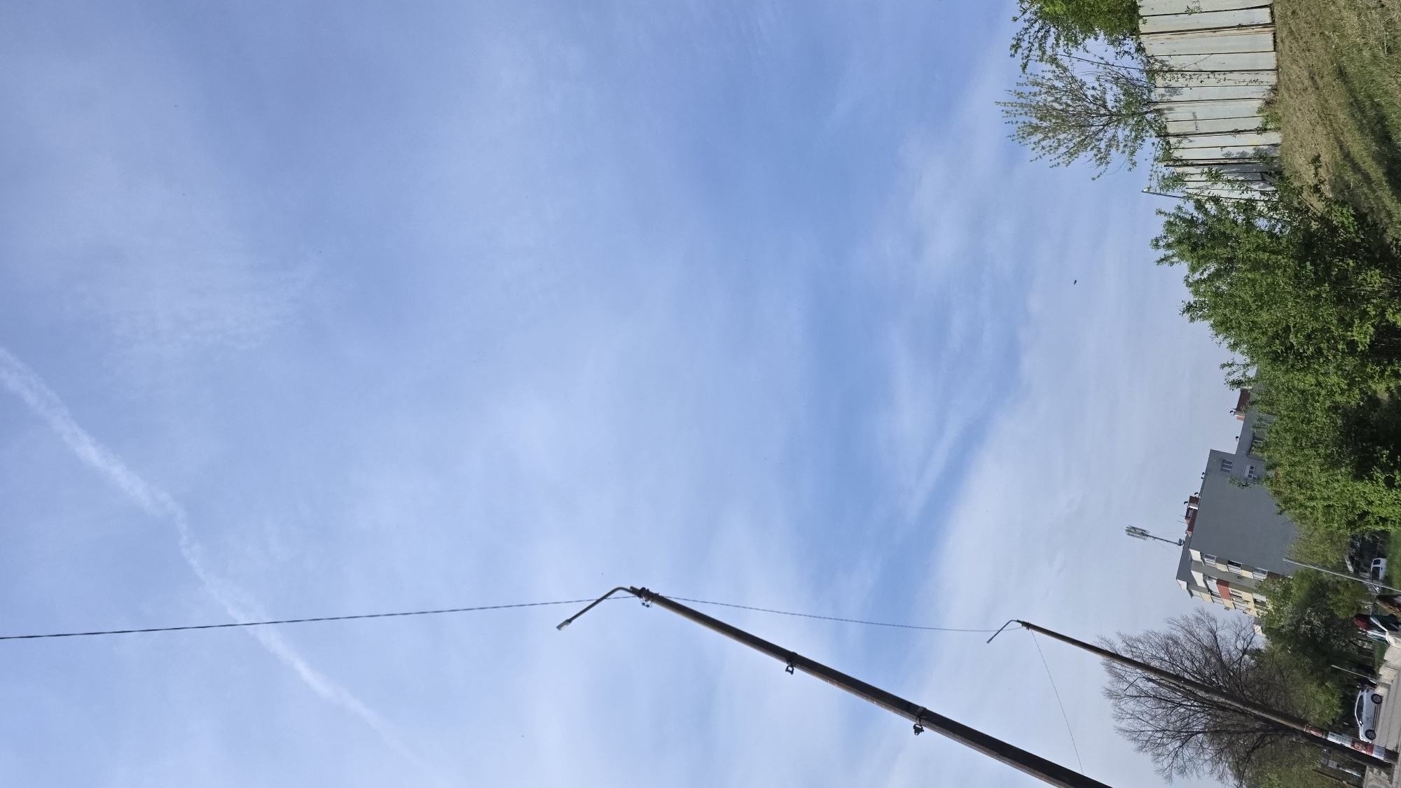 The image is a low-angle shot of a sky in Blagoevgrad, Bulgaria. The sky is predominantly blue with wispy clouds. An airplane contrail is visible in the upper left. Several utility poles are in the foreground, spanning diagonally from the bottom of the frame. Power lines extend horizontally across the sky.
In the background, a concrete wall borders a grassy area. Further back, there are buildings with trees surrounding them. Cars and other objects indicating street level activity are partially visible.
The image is oriented at an angle, causing the horizon to appear tilted.