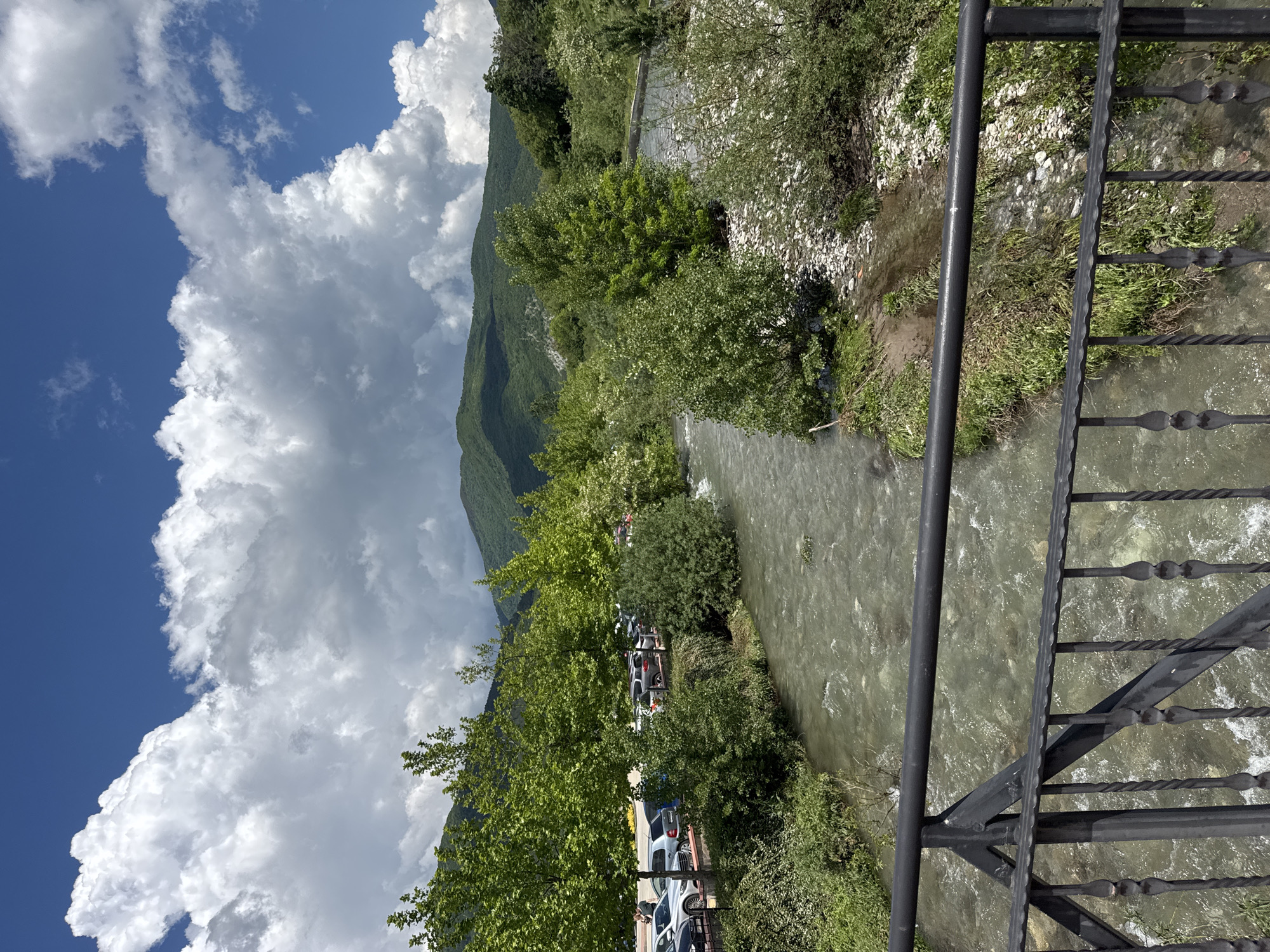 A medium-angle, full outdoor shot depicts a river scene from a bridge in Skobelevo, Bulgaria. The water runs vertically through the frame, with a railing visible on the right edge. The view encompasses a section of river and a parking area along its bank. On the left, a mountain looms, partially obscured by large, billowing white clouds against a blue sky. The river is lined with trees and other vegetation. Cars are parked in the parking area along the river's bank. The bridge's black metal railing is in the foreground, out of focus. The overall scene is naturalistic.