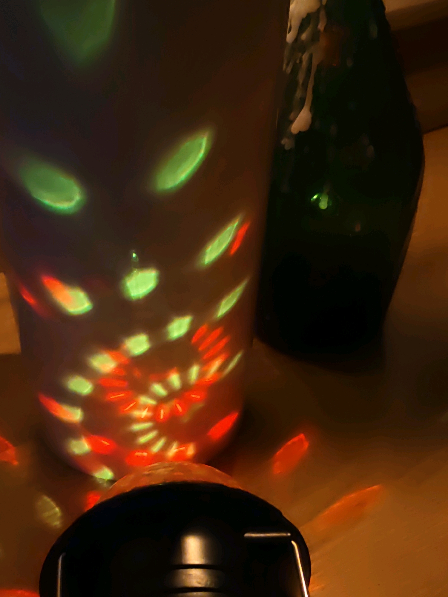 The image depicts a close-up, low-angle shot of a rotating disco ball projecting green and red lights onto a white cylindrical object and a dark green glass bottle. The disco ball is black and sits at the bottom of the frame, with its reflective surface partially visible. The white cylinder, likely a vase or a large cup, is positioned to the left and is illuminated by abstract patterns of light. To the right of the cylinder is the dark green bottle, which appears to have some foamy residue near its neck. The background is a wooden surface, possibly a table, and the overall lighting suggests a dimly lit indoor environment. The image does not contain any information that would allow for identification of a specific location in Rostov-on-Don, Russia.