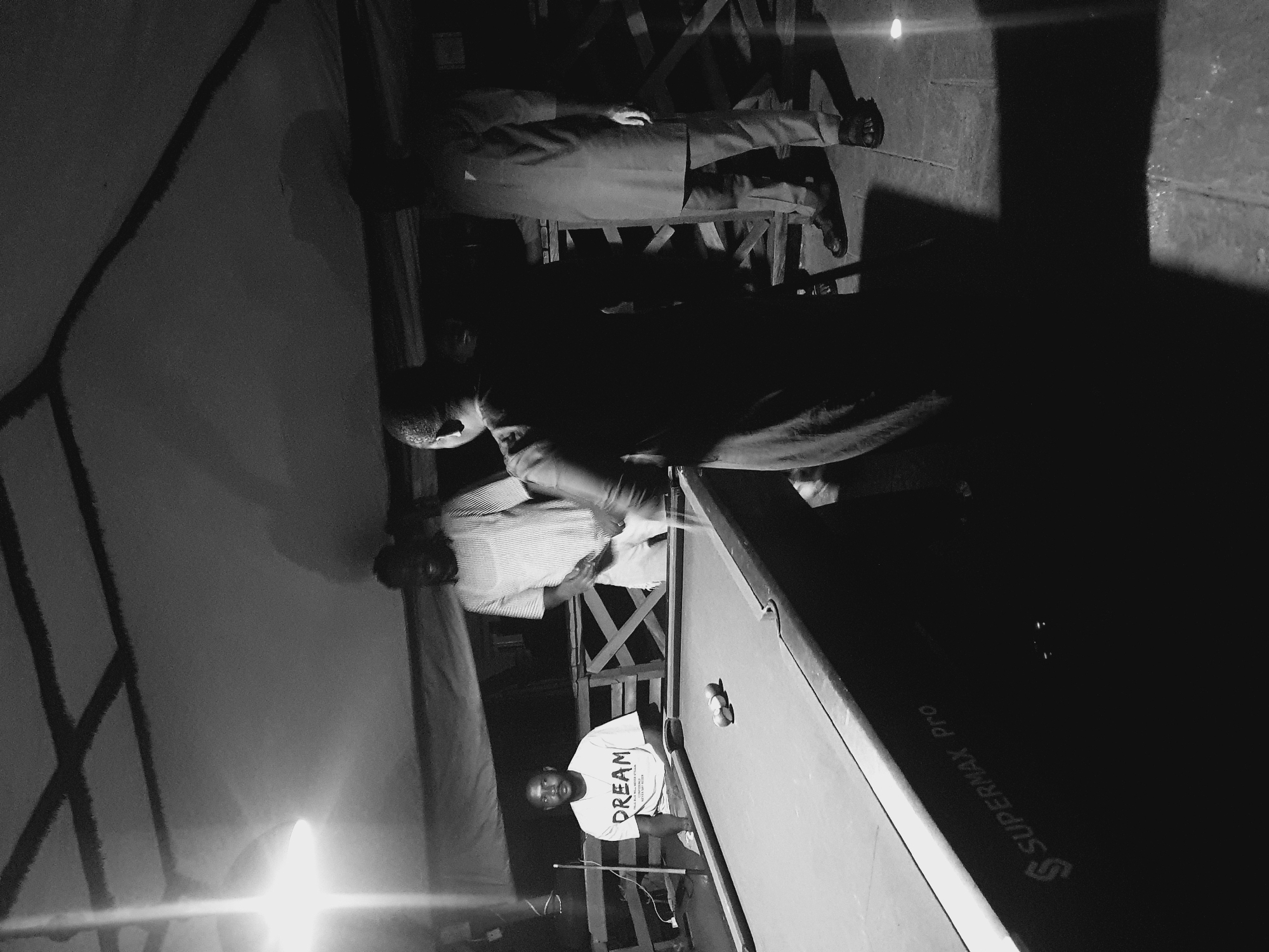 The black and white photograph captures a group of men gathered around a pool table in what appears to be an outdoor or semi-outdoor setting. The image is oriented sideways.
In the foreground, a man wearing a t-shirt with the word "DREAM" printed on it is positioned near the pool table, holding a pool cue. He is looking towards the camera. Three pool balls are visible on the table.
In the mid-ground, another person, possibly a man, is leaning over the pool table, engaged in the game. They are wearing a horizontally striped shirt. A third person, dressed in a suit, stands further back, their legs and feet visible.
The setting suggests a recreational area, possibly a bar or a gathering space, with a pool table as the central focus. The lighting is dim, casting long shadows, which might indicate it is nighttime or late evening. The presence of a large, tarp-like structure on the left side of the image could suggest an awning or temporary shelter.
The visible text on the pool table is "SUPERMAX PRO". The overall mood of the image is casual and focused on the game of pool. There are no immediate cues about the weather, but the lighting suggests a controlled environment. The location context of Lokoja, Nigeria, does not provide any specific visual elements within the image itself.