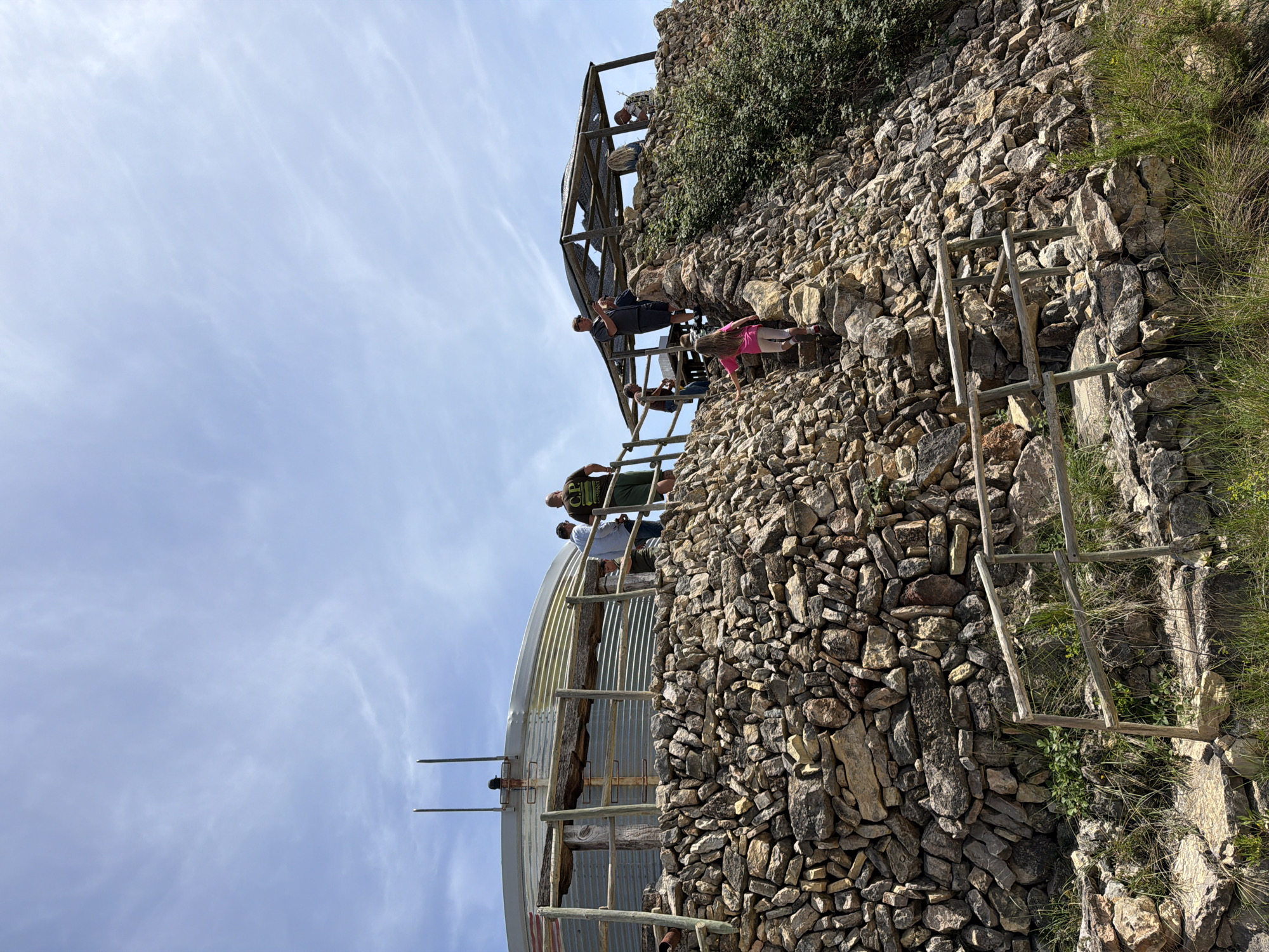 The image, taken in Bonastre, Spain, captures an outdoor scene featuring a group of people exploring an intricate stone wall construction, likely terracing or the ruins of an old building, set against a bright sky. The perspective looks upwards at the structures.
Several individuals are visible, including adults and possibly children, engaged in climbing and walking along the stone walls and integrated wooden structures. To the left, a large, curved structure with corrugated metal siding is partially seen, appearing to be built into the hillside. Above this, a group of people are gathered on a wooden platform or walkway. One man on this platform is wearing a dark green t-shirt with the text "C.P. COMPANY" clearly visible.
To the right, the stone construction continues upwards, characterized by meticulously stacked, irregular stones forming walls and steps. Wooden ladders or steps are incorporated into the stone pathways to facilitate movement up the steep incline. A person in a pink top and light trousers is seen among these stone steps. Lush green bushes and patches of dry grass grow around and on the stone structures, indicating a natural, somewhat overgrown environment.
The sky is a light blue with scattered, wispy white clouds, suggesting clear and sunny weather during the day. The overall activity appears to be recreational, possibly hiking or exploring a historical or natural site.