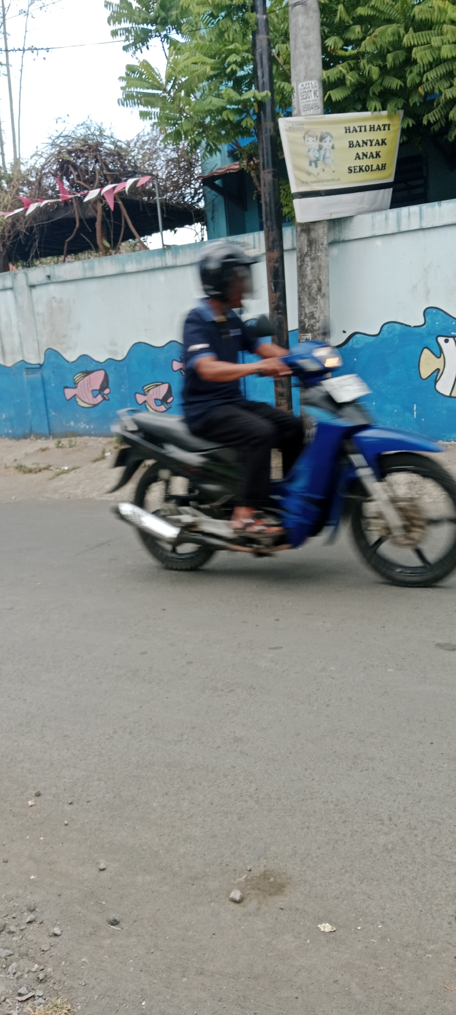 A person wearing a dark blue shirt, black pants, and a black helmet rides a blue motorcycle along a paved road. The motorcycle's wheels are blurred due to motion. In the background, a light blue wall is decorated with a mural of pink and white fish swimming in blue waves. A black pole stands to the right of the motorcycle, with a yellow banner attached that reads "HATI HATI BANYAK ANAK SEKOLAH," which translates to "Be Careful, Many School Children." The banner also features an illustration of two children. Green trees with dense foliage are visible above the wall and banner. The sky is overcast and appears white. The ground is asphalt, with some scattered small rocks and a small dark stain.