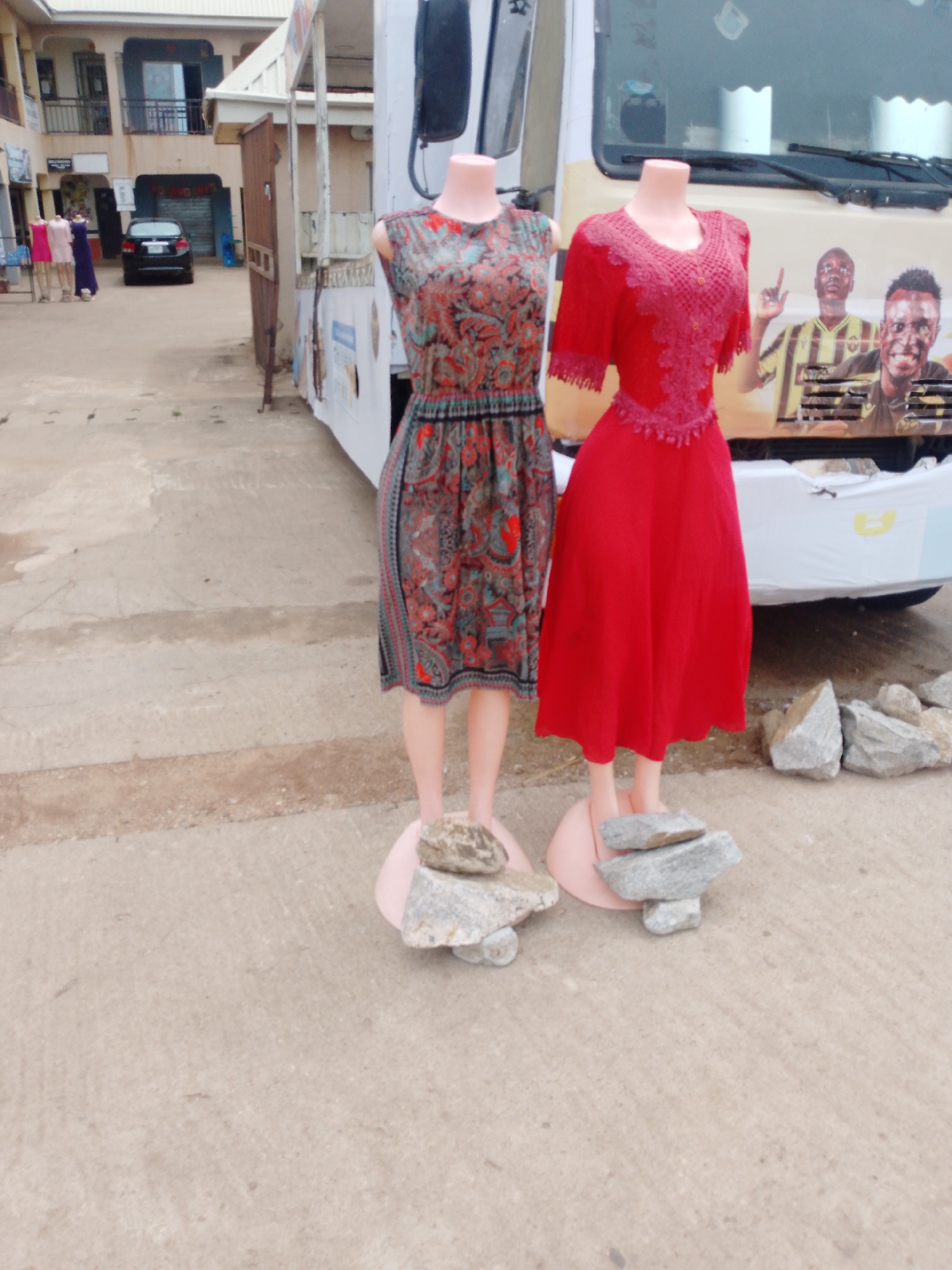 The scene is located in the city of Sabon Tasha, Nigeria, and features two headless mannequin torsos on pink bases, dressed in clothing. The mannequin on the left wears a paisley-patterned dress with a grey background, and the one on the right is dressed in a red dress with a lace top. Rocks are placed around the bases of the mannequins. The mannequins stand in front of a white van or truck with a photograph of people on the front. In the background, a building is visible. The street or concrete surface is lightly colored. In the far background, other people stand near a parked car.