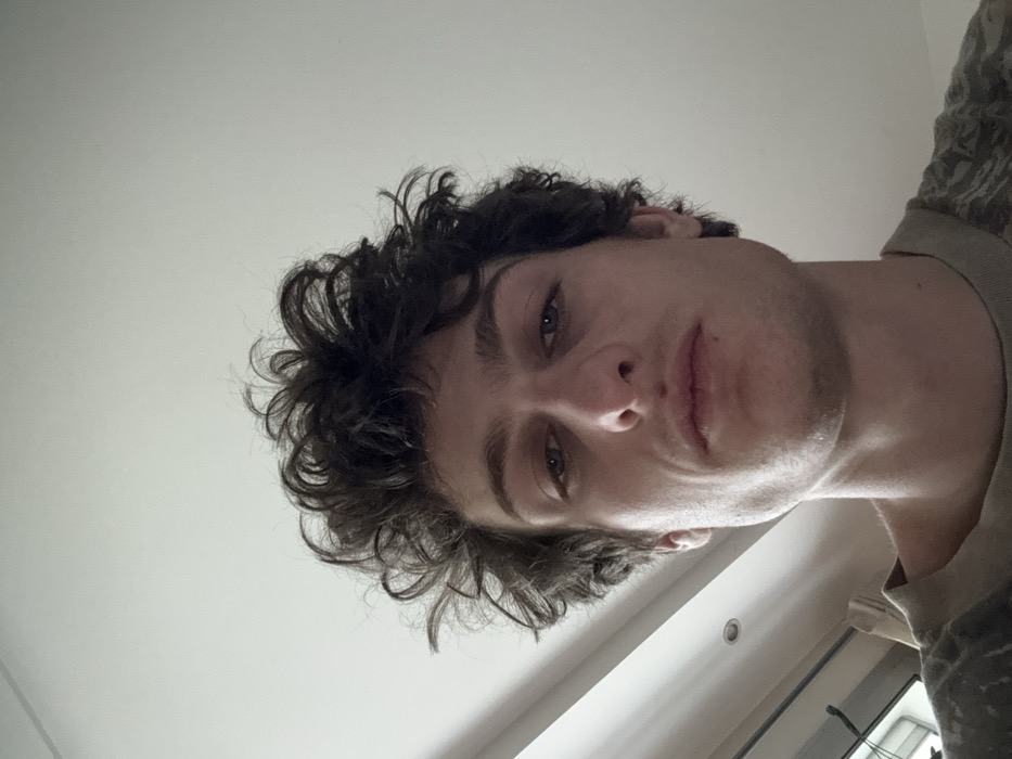 An individual identified as male is depicted in a close-up, head-and-shoulders portrait, centered in the frame. He has dark, curly hair and light-colored eyes. His facial expression is neutral, and he maintains a direct gaze towards the camera. He wears a light-colored t-shirt with a mottled or camouflage pattern. The background consists of a predominantly white, unadorned ceiling occupying the upper half of the image. A single recessed light fixture (downlight) is visible in the upper left quadrant of the background. In the lower left background, a partial view of an interior window or door frame, along with what appears to be a vertical blind or curtain, is discernible. The scene is situated indoors within the city of Vincennes, France. No specific actions or interactions are visible beyond the subject's pose for the photograph.