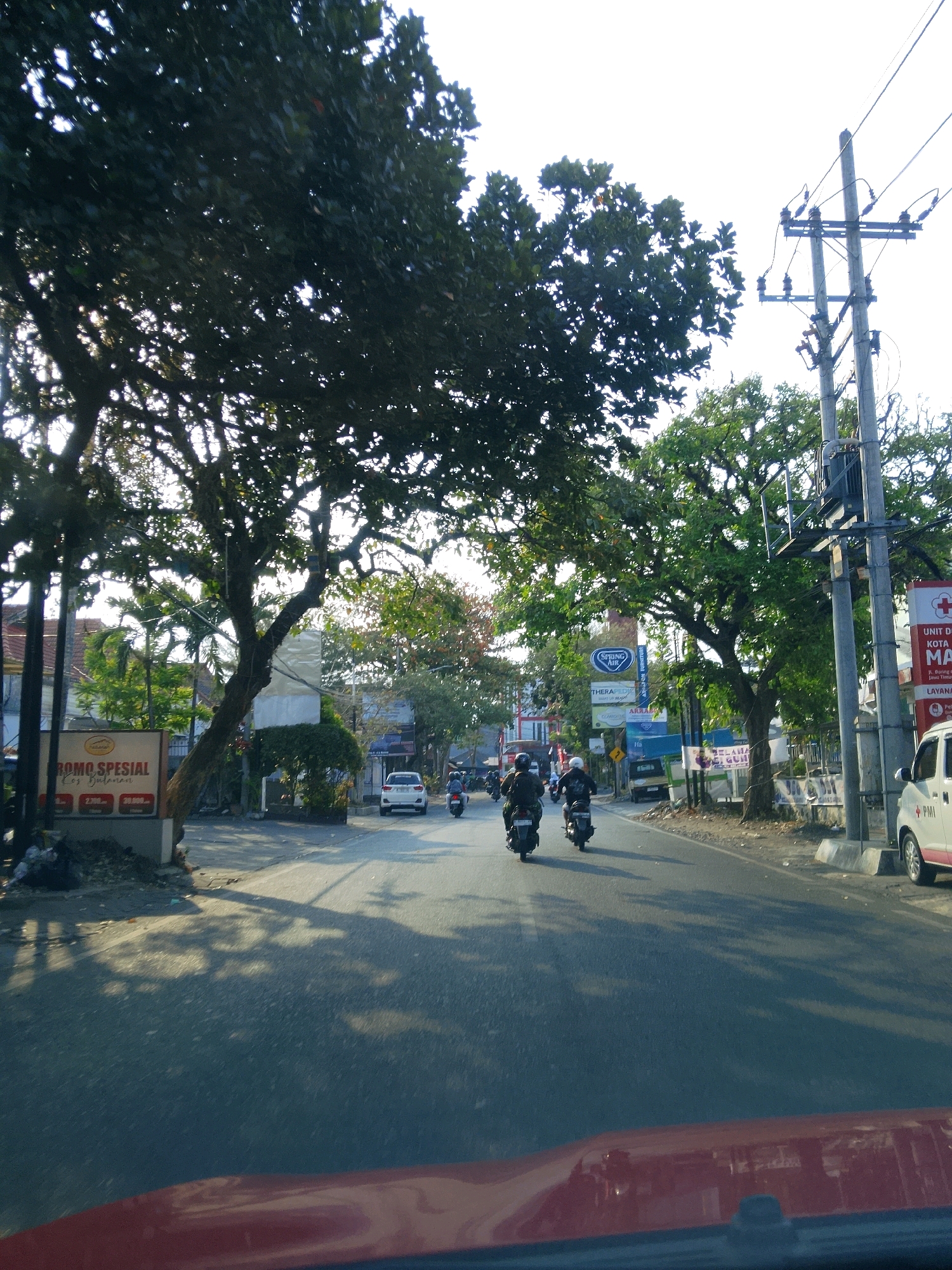 The image shows a street scene in Malang, Indonesia, likely during daylight hours given the bright sky and shadows. The perspective is from a vehicle, with the hood of a red car visible in the foreground.
The road is paved and appears to be two lanes wide. On the left side of the road, there is a large tree with dense foliage, casting shadows onto the road. A sign with Indonesian text, "PROMO SPESIAL," is visible near the base of this tree. Behind the tree, several buildings with tiled roofs are partially visible. Further down the road on the left, there are more trees and some street signs.
On the right side of the road, a utility pole with electrical equipment stands tall. More trees with green leaves line the side of the road. Several vehicles are present. Two motorcycles with riders are traveling away from the camera in the middle of the road. A white car is parked on the right side of the road. Another motorcycle is visible further down the road.
The environment appears to be an urban or semi-urban area. The sky is bright, suggesting it's daytime, and the light indicates the sun is likely in the sky. The overall impression is a typical street scene in a moderately busy Indonesian city.