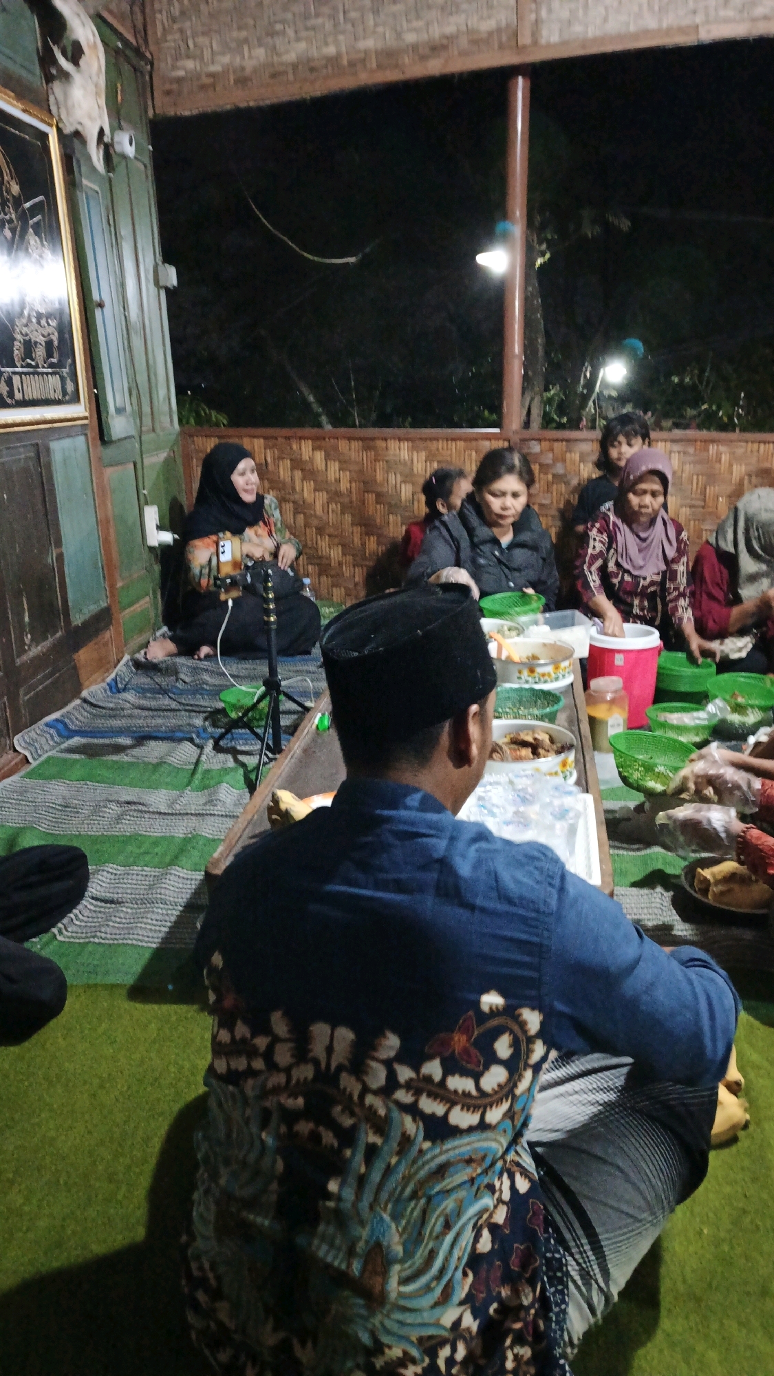 In Pojok, Indonesia, a group of people are gathered for a meal. The scene is set in an outdoor or semi-outdoor space with bamboo walls and a thatched roof. Several individuals, including women wearing hijabs, are seated on the floor around a low table laden with food. A man wearing a blue batik shirt and a black peci hat is in the foreground, his back to the camera, also seated on the floor. The lighting appears to be artificial, with some lamps visible in the background illuminating the dark night sky. The overall atmosphere suggests a communal gathering or a celebratory event.