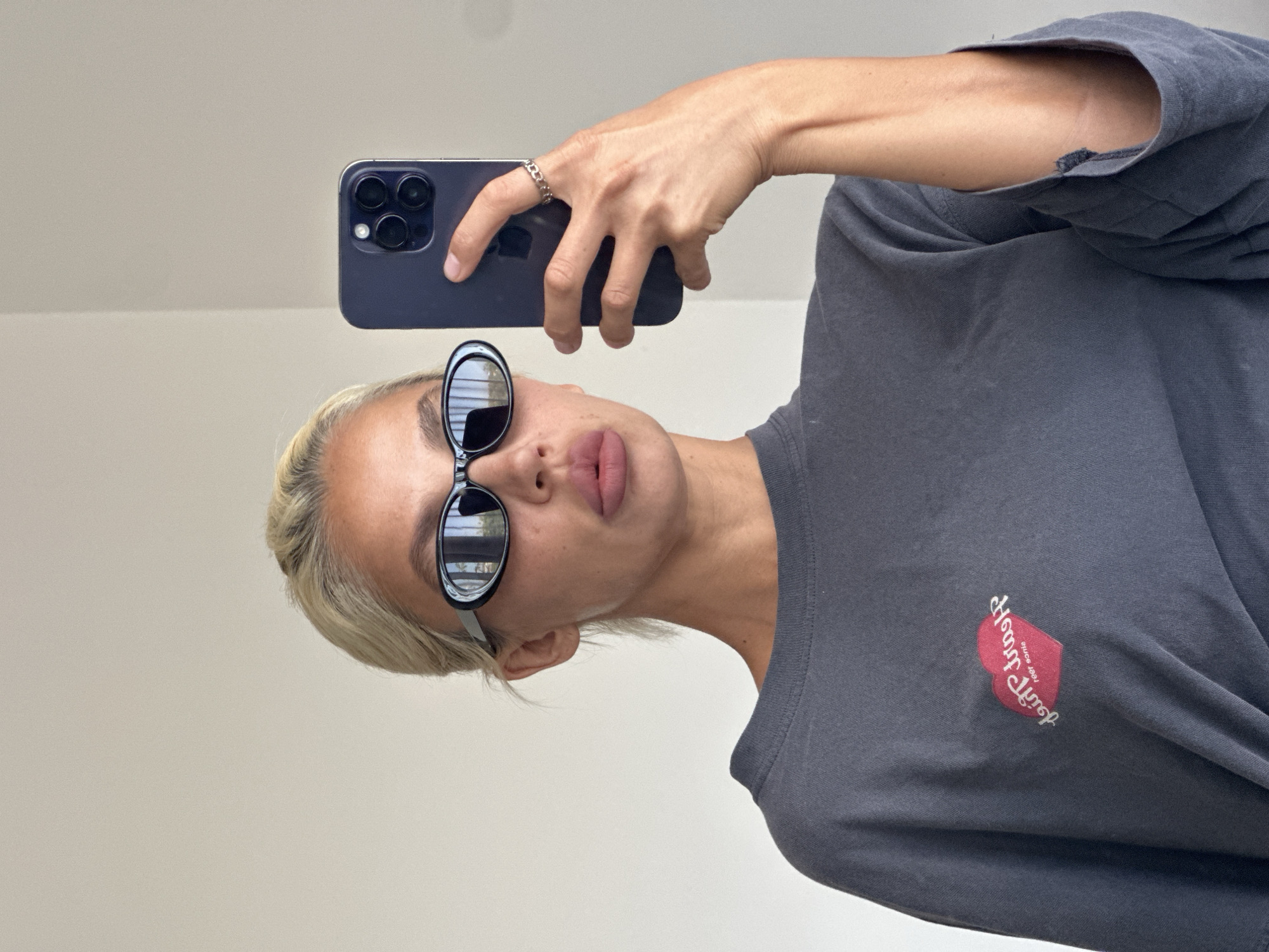 A blonde woman with full lips, wearing sunglasses and a dark grey t-shirt with a red heart logo, takes a selfie in a room with a light grey wall. She holds a dark purple smartphone in her right hand. The t-shirt has a small red heart graphic with white lettering on the left chest. A silver chain link ring is on her index finger. The image is cropped at the shoulders, obscuring the lower body and background details. The location is Kuta Selatan, Indonesia.