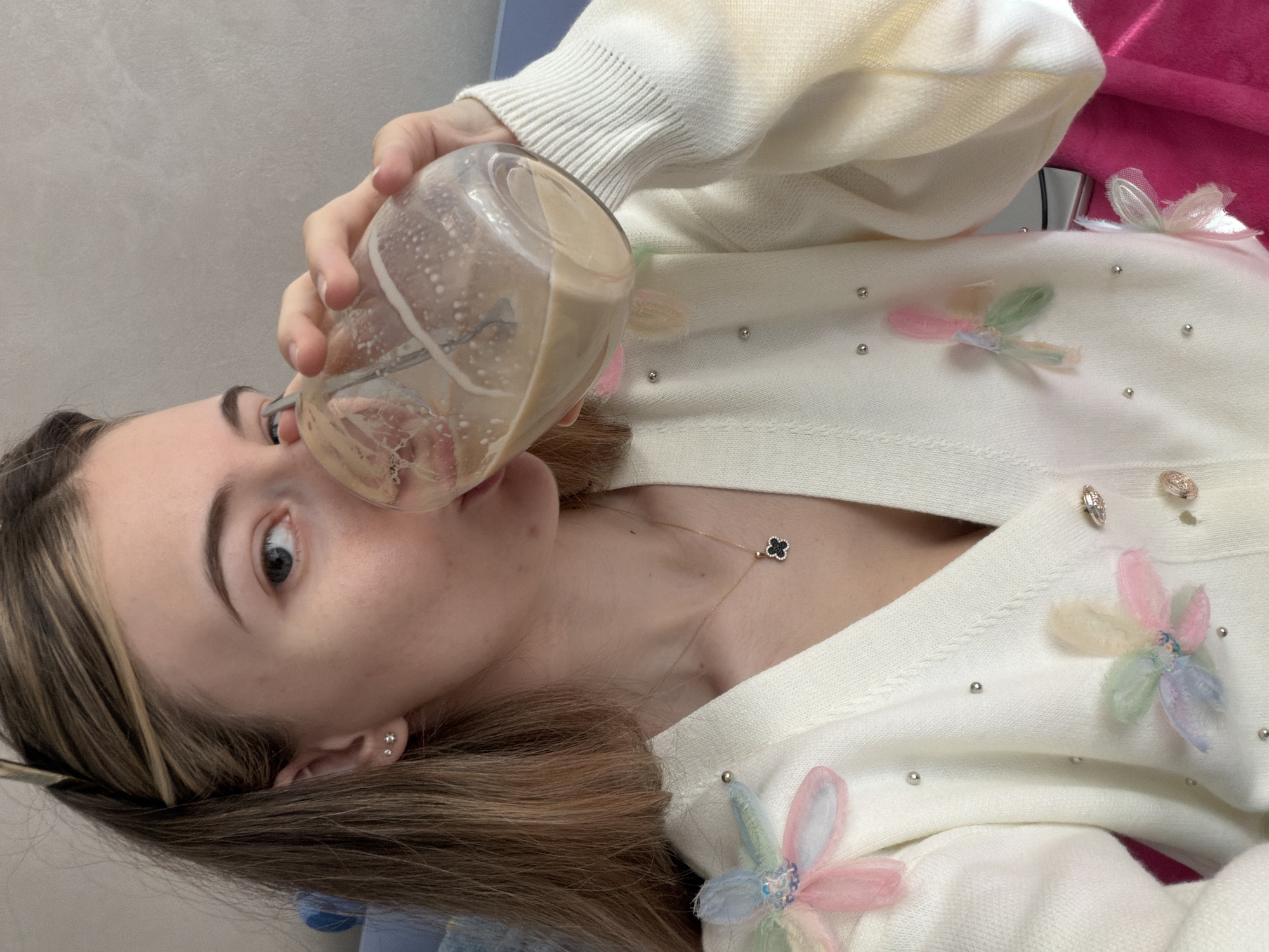 A woman is drinking from a glass jar. The jar is almost empty and contains a light brown liquid with bubbles. The woman has brown hair with blonde highlights, fair skin, and is wearing earrings. She is wearing a white cardigan sweater with floral embellishments and small silver beads. She is also wearing a gold necklace with a clover pendant. The background appears to be a plain wall, likely indoors. There is no discernible evidence to associate this image with Plovdiv, Bulgaria.