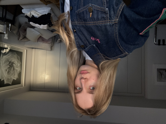 A young adult female with long, wavy blonde hair and light eyes is positioned in the foreground, facing slightly left of the camera, looking directly forward. She wears a dark denim jacket featuring pink "S&G" embroidery on the right chest and a green and pink patch on the upper left sleeve, over a white garment. The indoor background displays white paneled walls or closet doors in the center-right, illuminated by a light source from the right. To the left, a framed, light-colored abstract artwork hangs on a white wall, partially obscuring a metallic object below. To the right, a light-colored sofa is visible, covered with numerous pillows of various grey and pink hues, atop which a small pile of black and light-colored clothing rests. Adjacent to the sofa, a dark-colored side table holds a white vase containing white orchid flowers and several small containers. Above the sofa and table, a large framed artwork depicting a layered white flower against a dark background hangs on the wall. No specific visual cues identify the location as Mexico City, Mexico, within this interior scene.