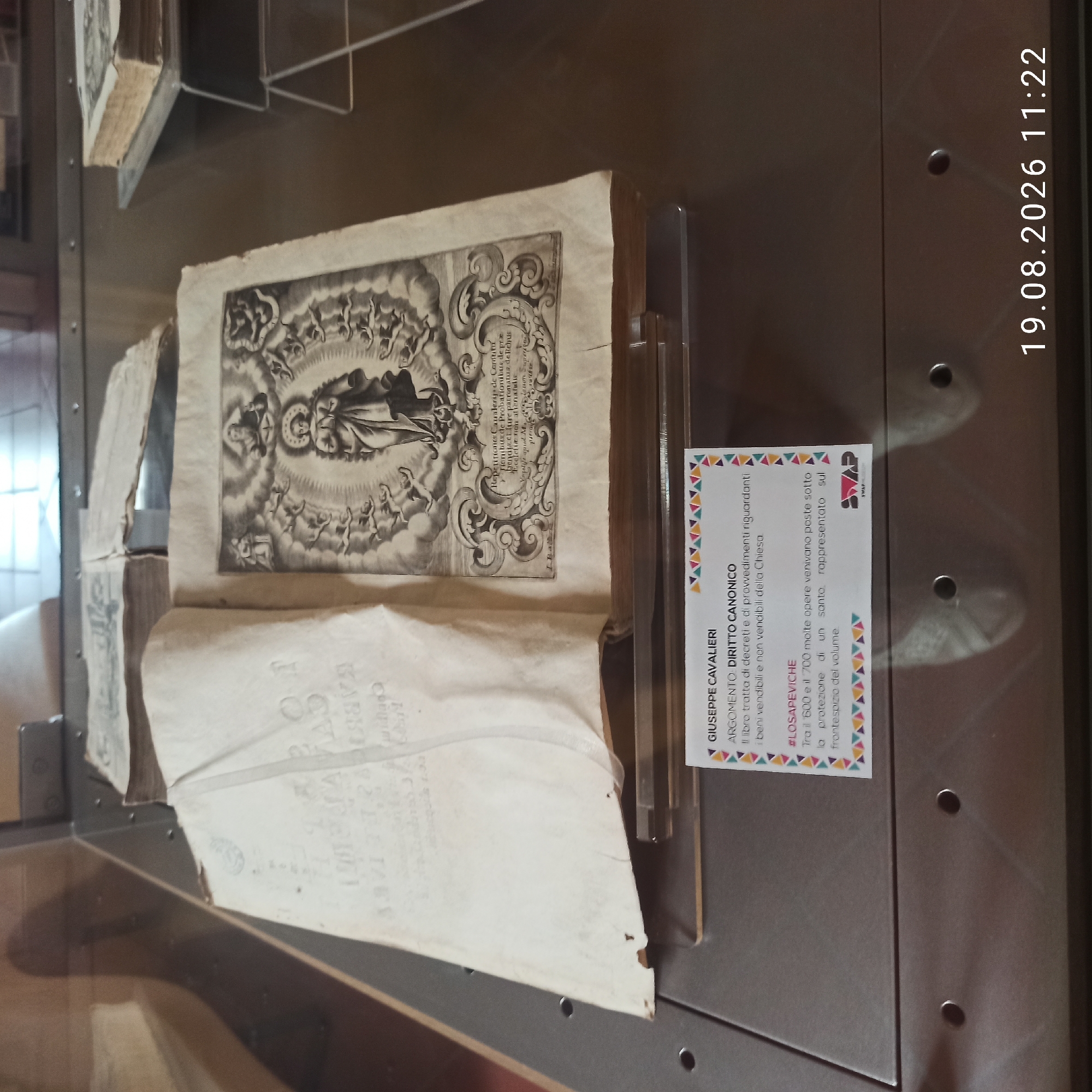 The image depicts an open, aged book displayed within a glass case, likely in a museum or archive in Ruvo di Puglia, Italy, based on the context provided by the information card. The left page of the book is largely blank, showing signs of wear and a watermark. The right page features an intricate engraving of a religious scene, possibly the Madonna surrounded by cherubs and other figures. Below the engraving, there is Latin text. A descriptive card placed in front of the book provides information in Italian. The card identifies the book as belonging to Giuseppe Cavalieri, with the subject being "DIRITTO CANONICO" (Canon Law). It further states that the book deals with decrees and provisions concerning the Church's movable and immovable property. A section titled "#LOSAPEVICHE" (Did you know) mentions that between the 1600s and 1700s, many works were placed under the protection of a saint represented on the frontispiece of the volume. The background suggests an indoor environment with muted lighting. The bottom right corner of the image shows a timestamp "19.08.2026 11:22".