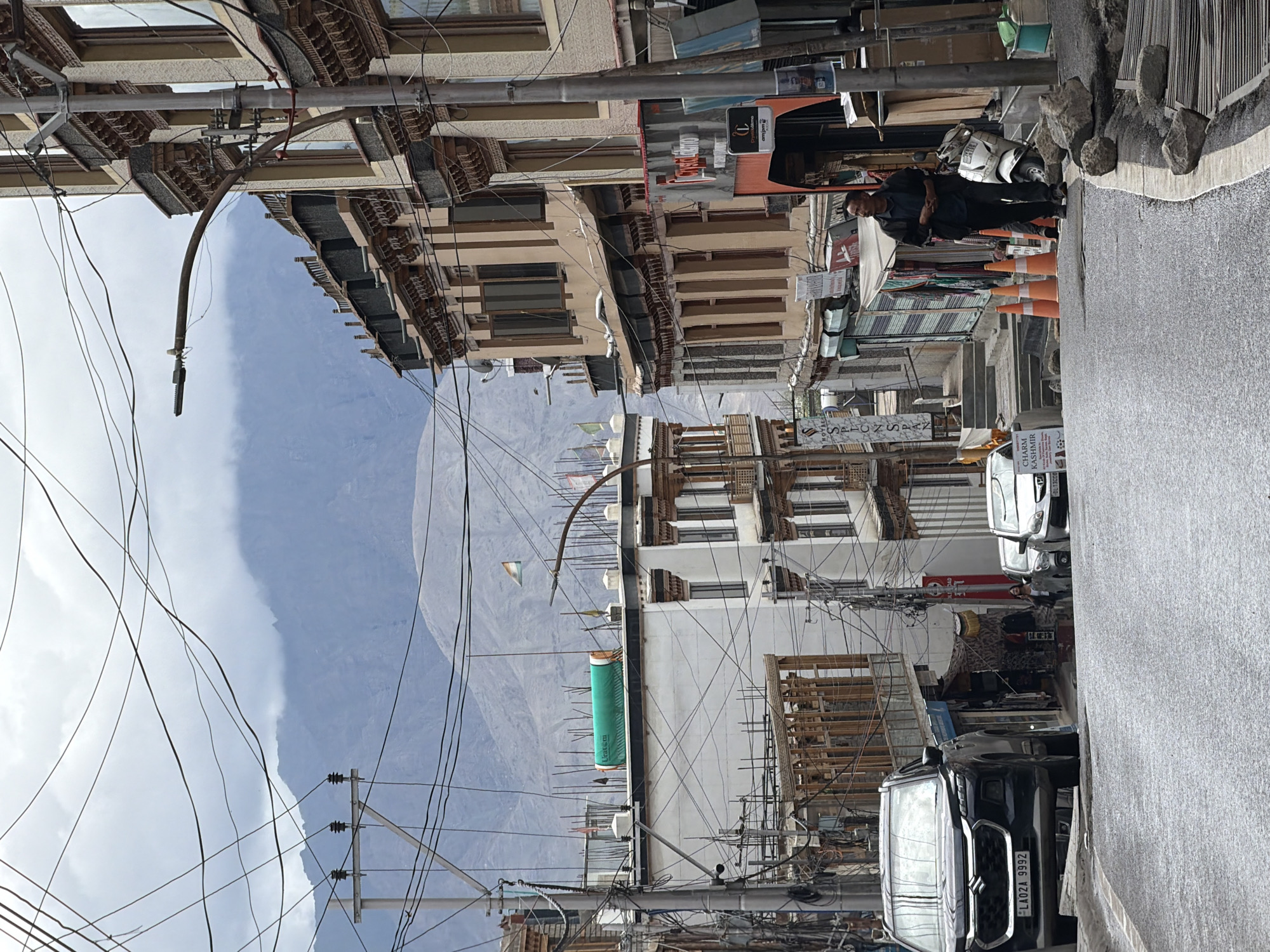 The image shows a street scene in Leh, India, with traditional Ladakhi architecture. A black SUV is parked on the left side of the road, facing uphill. In the background, mountains are visible under a cloudy sky. The street is lined with multi-story buildings, some with balconies and intricate wooden details. There are numerous power lines crisscrossing the sky. A man is walking on the right side of the street, dressed in dark clothing. A white scooter is parked nearby. Several orange traffic cones are placed on the side of the road. Signs for shops are visible on the buildings. The road surface appears to be wet, suggesting recent rain.