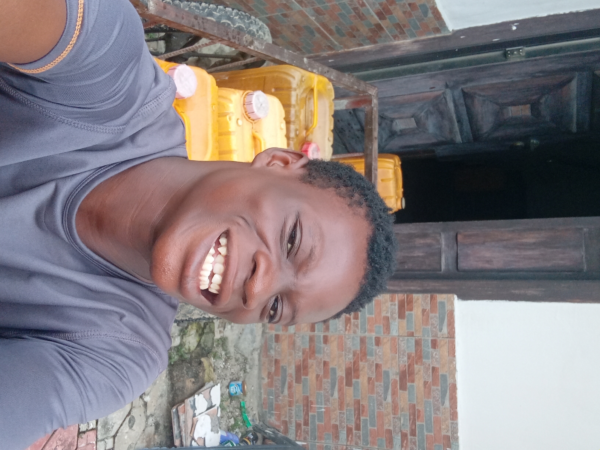 A young Black male is taking a selfie in an outdoor setting. He is wearing a dark gray t-shirt and is smiling widely, revealing his teeth. Behind him, there are several large yellow plastic containers stacked on a metal cart. The background also shows a brick wall and a wooden door. The ground appears to be paved with bricks and stones, with some debris scattered around. The overall scene suggests a storage or loading area, possibly in a residential or commercial district.
