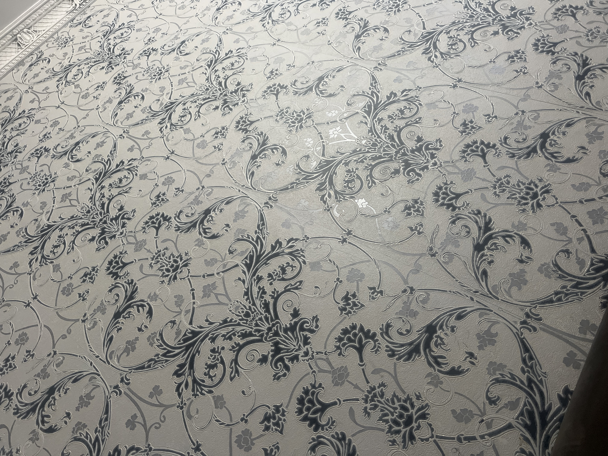 The image shows a close-up view of a textured wallpaper with a damask pattern. The wallpaper has a light beige background with a repeating design of dark gray and silver floral and vine motifs. The pattern is intricate and appears to be embossed, giving it a raised texture. The lighting suggests the wallpaper is on an interior wall, possibly in a home or a public building. There is a dark, curved object in the bottom left corner of the image, which could be a piece of furniture or trim. The scene is static and does not depict any specific activity or interaction. There is no visible indication of the city of Akhalkalaki, Georgia, or any other specific location.