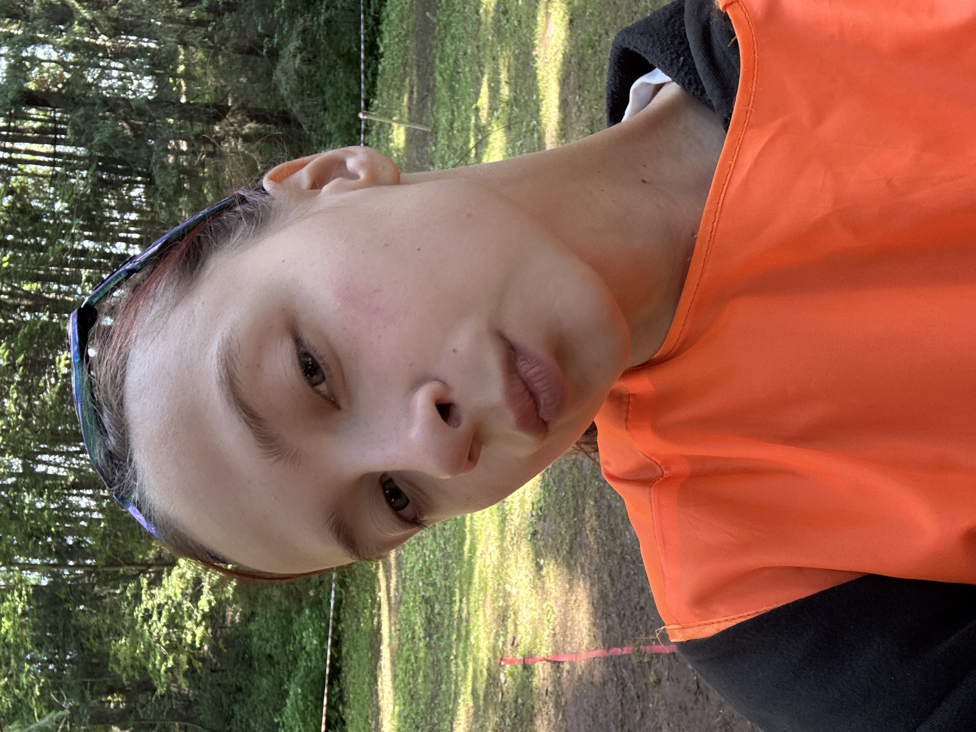 The media file shows a close-up, Dutch angle shot of a young person outdoors. The subject has light skin, light brown hair, and is wearing an orange vest over a black garment. They are also wearing blue-tinted eyeglasses on their head. The background is blurred but appears to be a natural, wooded environment with green grass and trees, suggesting a park or forest setting. Sunlight is filtering through the trees, creating dappled lighting. The time of day appears to be daytime, likely late morning or afternoon, given the brightness and the shadows. The person's expression is neutral or slightly contemplative. There are no visible text elements in the image. The location context provided is Сергино, Russia. The scene suggests an outdoor activity or event, possibly a sporting event or a casual outing in nature, given the vest and the outdoor setting.
