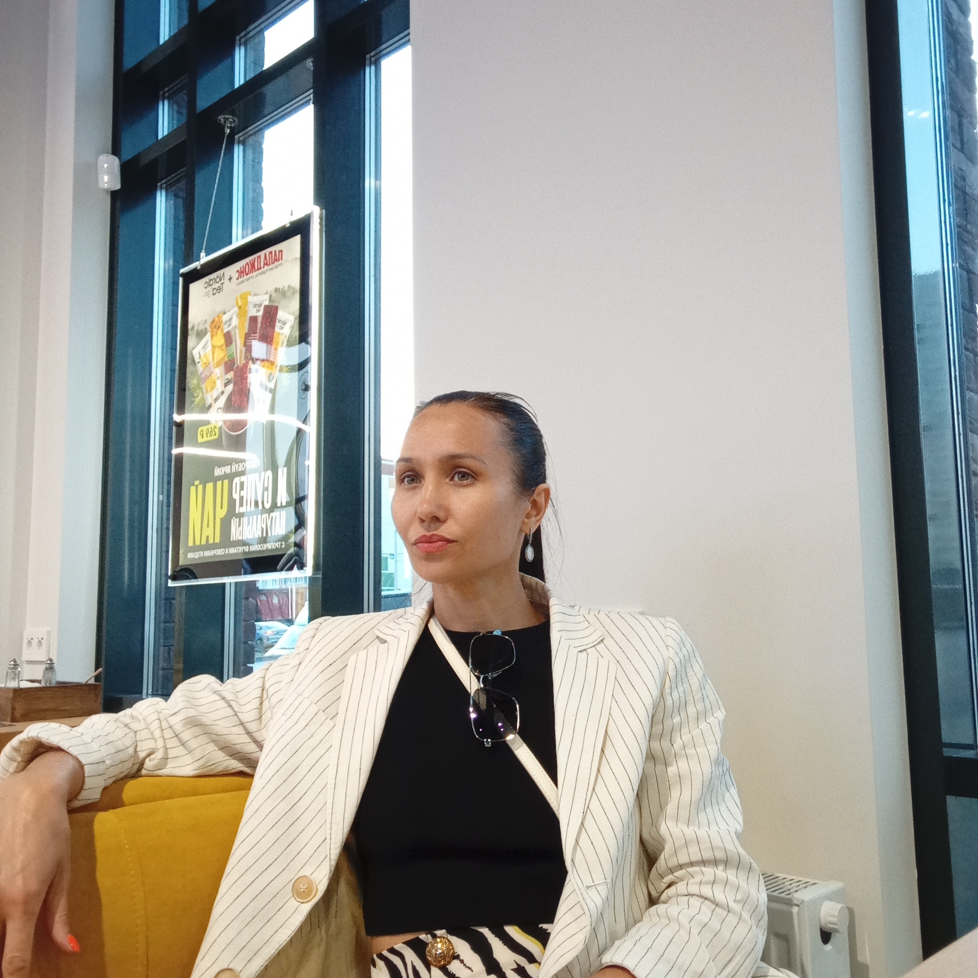 A woman sits on a yellow couch in front of a window with a poster hanging on it. She is wearing a white pinstripe blazer over a black top and a patterned skirt. Sunglasses are hanging from the neckline of her top. The poster on the window displays text in Cyrillic and images of tea packages. A white radiator is visible to the right of the woman. The room has white walls and dark window frames.