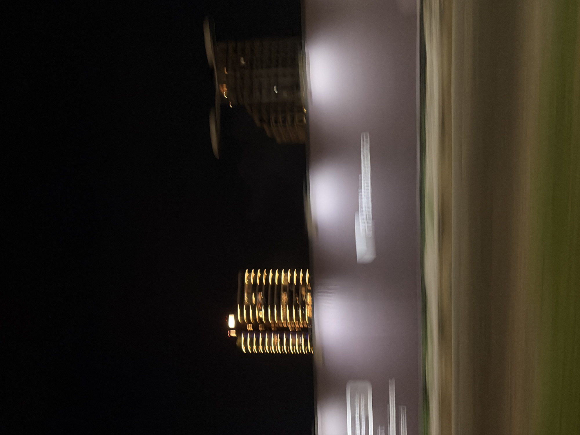 The image shows a nighttime scene likely in Ikoyi, Nigeria, with blurred lights suggesting motion. Buildings with illuminated windows are visible against a dark sky. A light-colored, possibly green or beige, elongated area extends across the frame, possibly representing a wall or facade. Signs or text are present in a lighter hue on a dark surface.