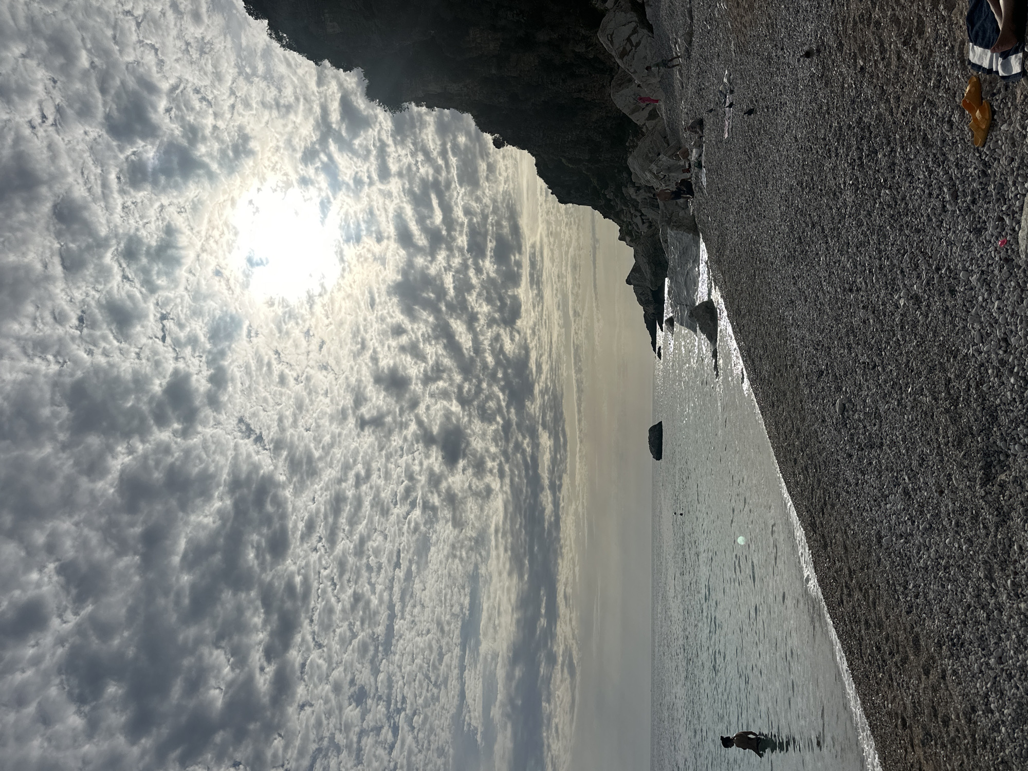 The image captures a detailed view of a pebbly beach in Mirište, Montenegro, under a sky largely covered by textured altocumulus clouds. The sun, partially obscured but brightly visible through a break in the clouds, is positioned in the upper-middle right quadrant.
The scene features a wide expanse of small, gray and white pebbles forming the beach in the foreground and extending towards the mid-ground. To the left, the calm, greenish-blue sea meets the shore with gentle waves. One individual is partially submerged in the water on the far left, facing away from the viewer. Further out in the water, a large, dark rock formation is visible.
A prominent, dark, rocky cliff face dominates the right side of the image, extending from the middle to the upper right. White boulders are scattered at the base of this cliff and along the water's edge. A small group of approximately 3-4 individuals are visible sitting or lounging on the pebbles near the base of the cliff. One person in a pink garment is distinguishable near the white rocks, and another, possibly a child, stands closer to the cliff face.
In the bottom right corner of the foreground, a pair of bright yellow flip-flops rests on the pebbles next to a blue and white striped towel. Two small, dark birds are also visible on the pebbled beach surface near these items. The overall lighting suggests either early morning or late afternoon, with the sun's rays diffused by the extensive cloud cover.