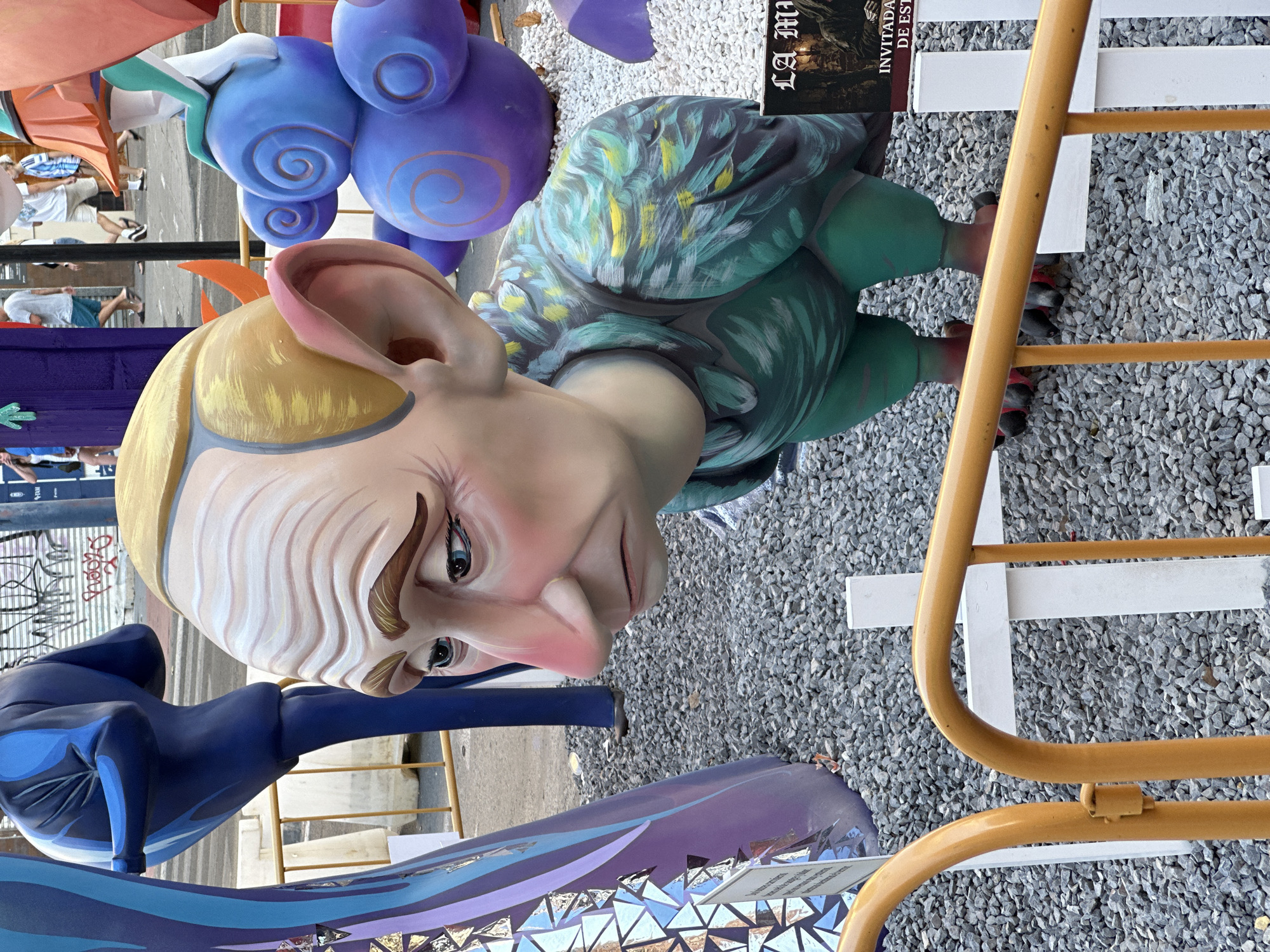 The image depicts a street scene featuring elaborate, oversized figurative sculptures, typical of a Fallas festival, likely in Alicante, Spain. The primary subject is a large, stylized head of a person with exaggerated features, including large ears, prominent nose, and wrinkled skin rendered in pale tones with rosy cheeks. The figure's blond hair is painted, and its eyes are a piercing blue.
Behind and to the left of the main head, other colorful sculptures are visible. These include a large, coiled, purple shape resembling a cloud or a character, and parts of other figures in orange and blue. The sculpture in the foreground, on the right, is a torso covered in a green and yellow painted patterned fabric, with red accents on its feet.
The sculptures are set against a backdrop of gravel and a white picket fence, with a bright yellow metal railing in the foreground. In the background, parts of other figures and structures can be seen, as well as some people observing the displays. The lighting suggests it is daytime, possibly sunny, casting shadows.
A small sign is visible attached to the green sculpture, featuring some text in what appears to be a gothic font. The visible text reads "LA MAN" followed by "INVITADA DE EST". The overall impression is one of a festive, public display of art and cultural tradition.