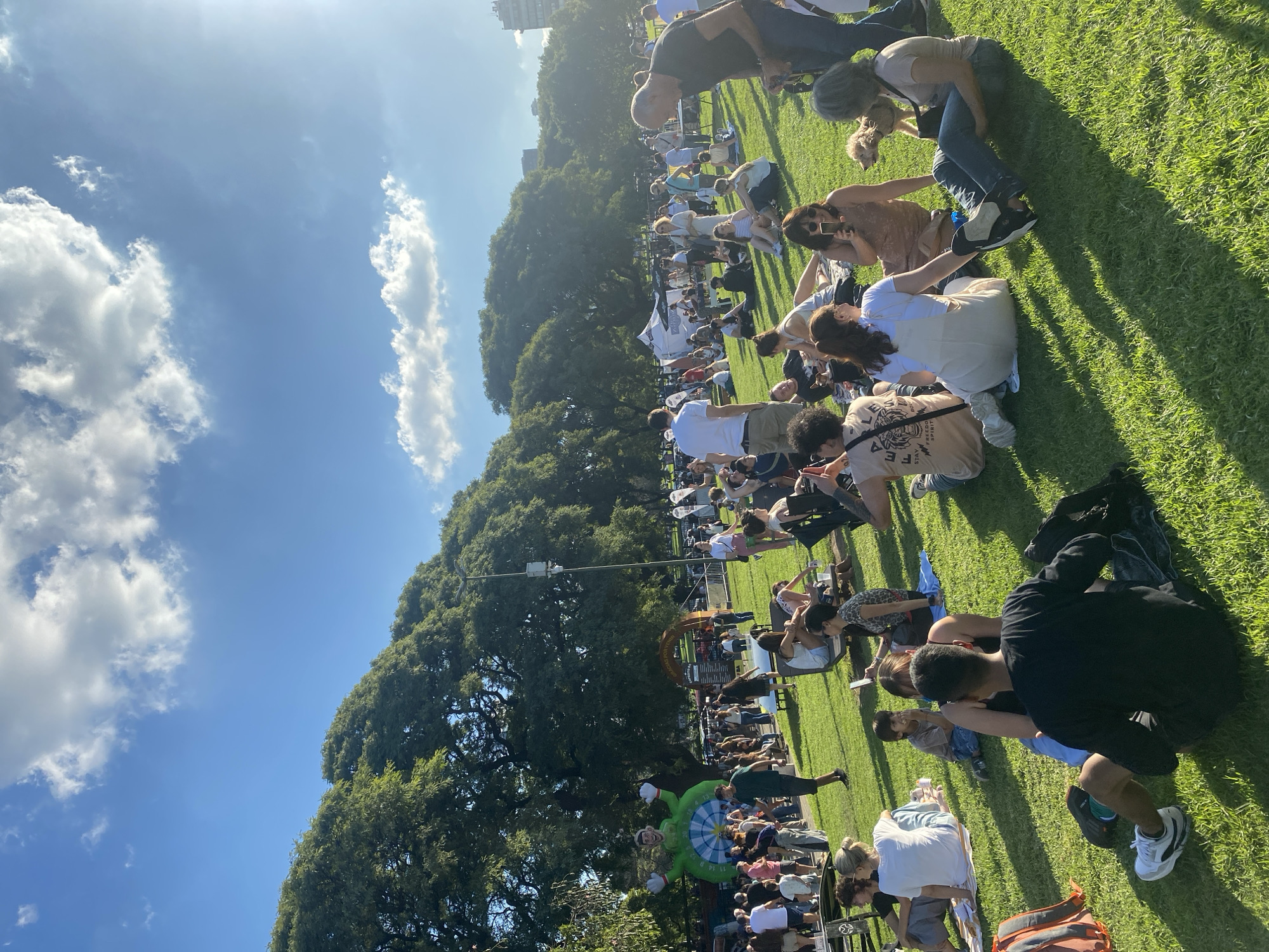 An elevated, long shot presents a crowd of diverse individuals spread across a large, manicured green space under a bright, slightly hazy sky with scattered clouds. The locale appears to be an outdoor event, possibly a fair or festival, given the presence of tents and an inflated character with a dartboard design. The crowd is primarily made up of people sitting or crouching on the grass. Some individuals are engaged in activities, such as taking photographs or talking to one another. A line of mature trees borders the green space, partially obscuring buildings in the distance. The location could possibly be in the city of Buenos Aires, Argentina.