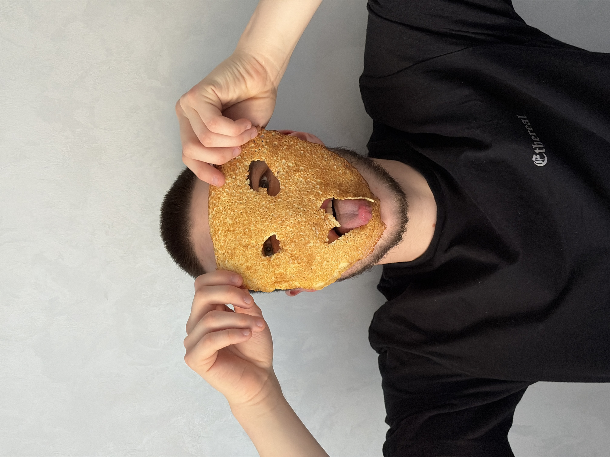 A man is holding a pancake with holes in it over his face, obscuring it. His tongue is sticking out through one of the holes. He is wearing a black long-sleeved shirt. The background is a light gray surface.
There is no indication of the location being in the city of Sarapul, Russia.