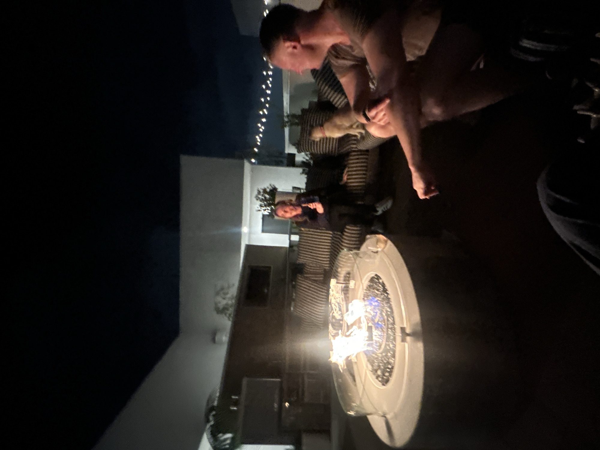 The image depicts an outdoor nighttime gathering on a modern rooftop or patio, potentially situated in Los Angeles, United States of America. A prominent circular gas fire pit, featuring a glass wind guard and emitting bright flames, occupies the central foreground, serving as the primary light source.
Two human subjects and one canine subject are present. To the left, a female with light hair is seated on a black and white striped outdoor sofa. She wears dark clothing and light-colored athletic shoes, holds an unidentified object near her chest, and appears to be speaking or laughing while looking towards the right. Adjacent to her on the same sofa, a light-colored dog, possibly a Labrador Retriever, sits attentively, facing left. In the right foreground, a male with short light-colored hair is seated on the ground or a low surface, positioned in profile facing left. He wears a short-sleeved top and dark shorts, with his legs bent and his right arm resting on his knee, engaging with the female subject.
The background features contemporary architecture with stark white walls and flat rooflines. A dark, rectangular structure is visible behind the fire pit, incorporating what appears to be an unlit television screen. Potted plants are strategically placed. String lights with numerous small, warm-toned bulbs are strung across the upper right portion of the frame, providing additional ambient illumination against the dark night sky. The ground surface is dark and unidentifiable. The scene conveys a relaxed social interaction.