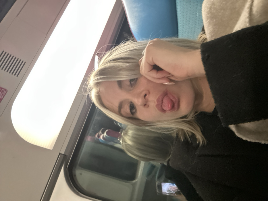A woman with blond hair poses in what seems to be a train. She has one hand near her forehead, her lips are puckered, and she is sticking out her tongue. She's wearing a dark jacket and a ring on her left ring finger. A window with a reflection is behind her and a bright light is above. The image is slightly angled.