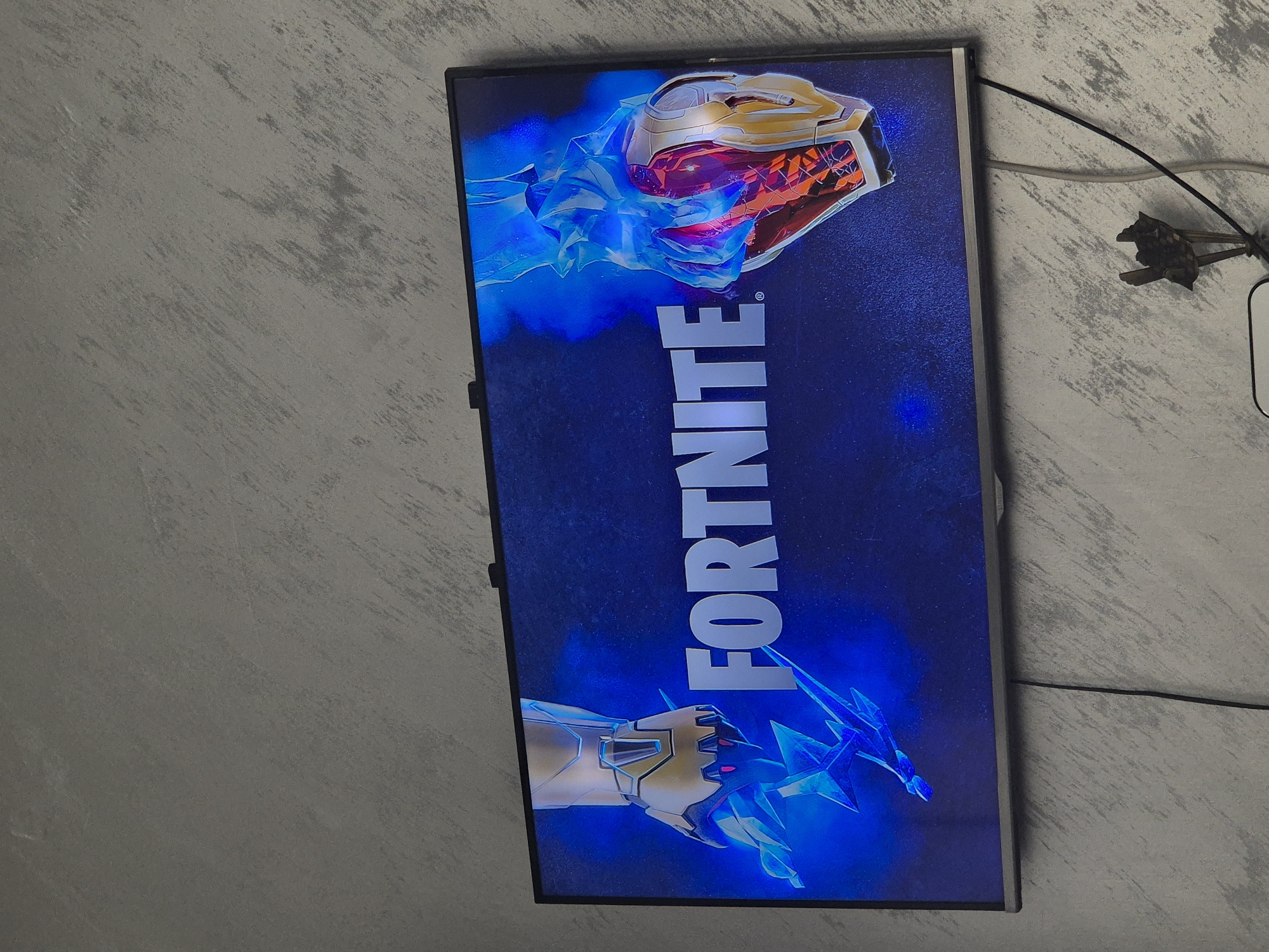 A monitor displaying the game "Fortnite" is mounted on a textured wall. The screen shows a helmeted figure at the top and a gloved hand at the bottom, both surrounded by blue energy. Cords hang to the right of the screen. The location is unknown.